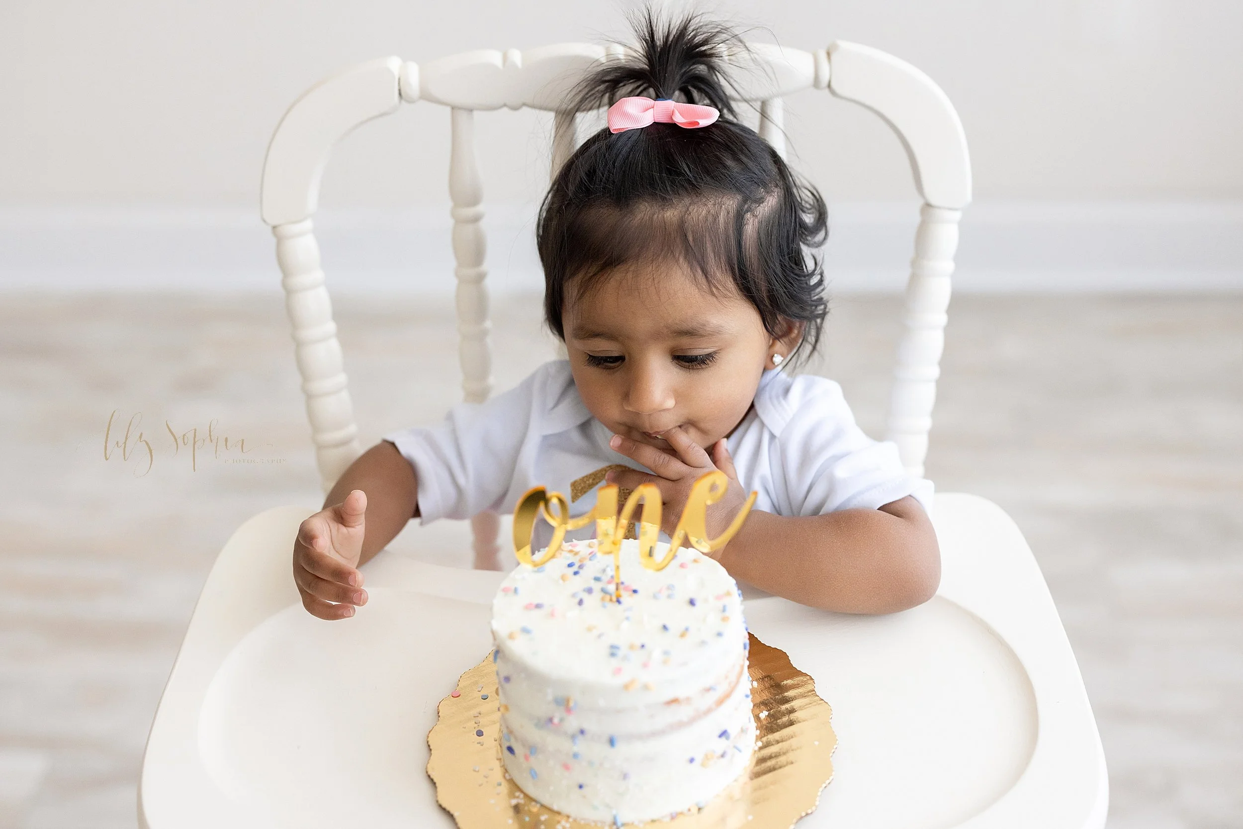  Overhead shot of a one year old little girl celebrating her first birthday with a smash cake on the tray of the antique highchair she is sitting in with her finger giving her the first taste of the icing on her cake taken in a natural light studio n