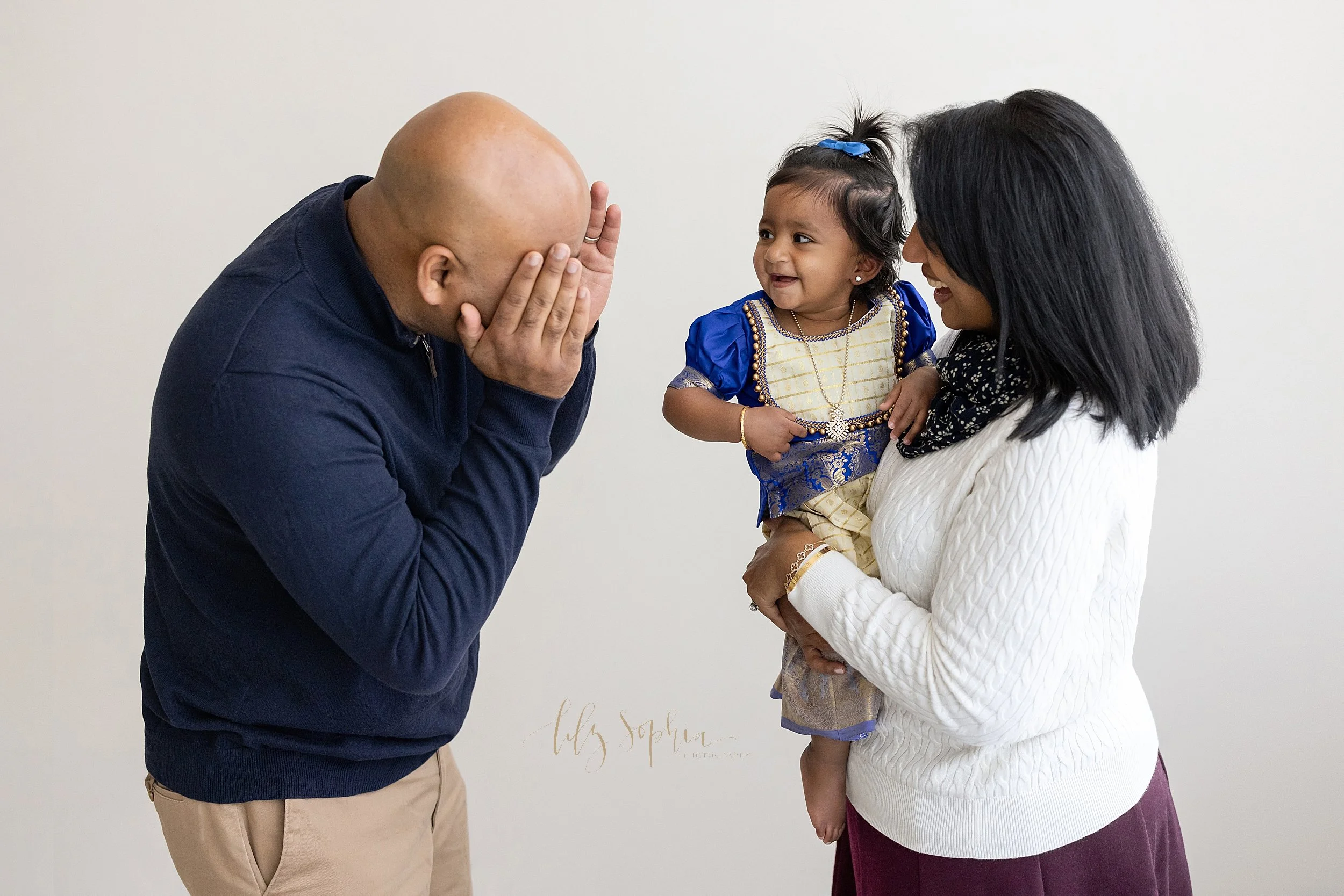  Family portrait of an Indian mother holding her one year old daughter in her arms as she stands facing her husband who is playing peek-a-boo with his daughter taken near Buckhead in Atlanta in a natural light studio. 