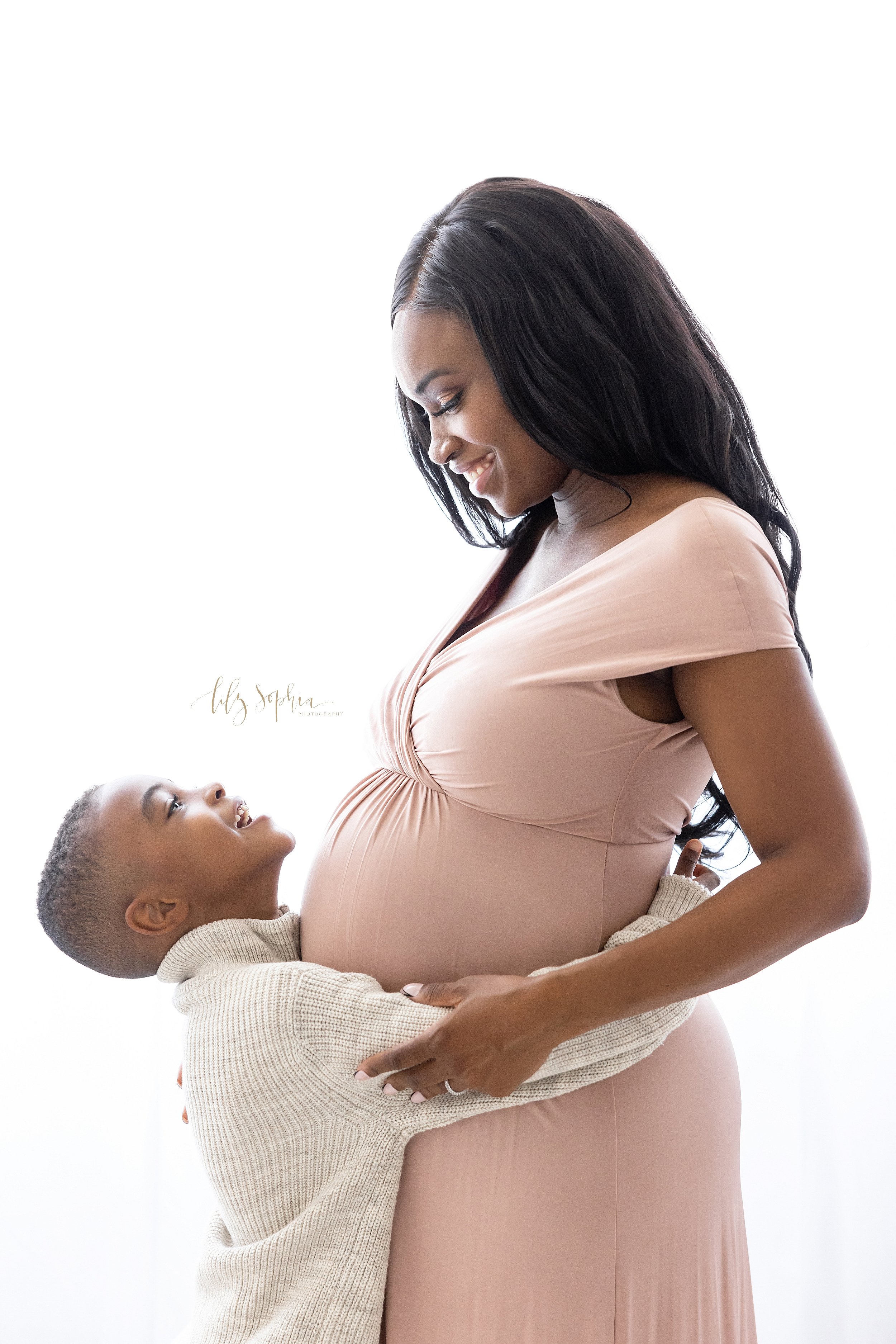  Family maternity portrait of an African-American expectant mother and her young son as they stand in a natural light studio with the son wrapping his arms around his mom’s waist and the two of them look at one another taken near Oakhurst in Atlanta,