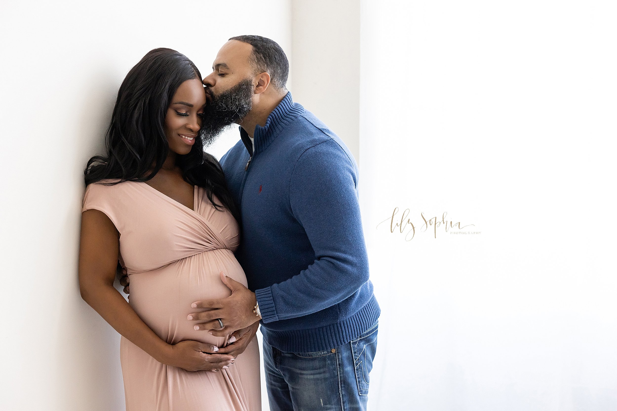  Maternity photo of an African-American father kissing the forehead of his wife as he wraps his right hand around her back and places his left hand on his child in utero while the couple stand in a natural light studio near Poncey Highlands in Atlant