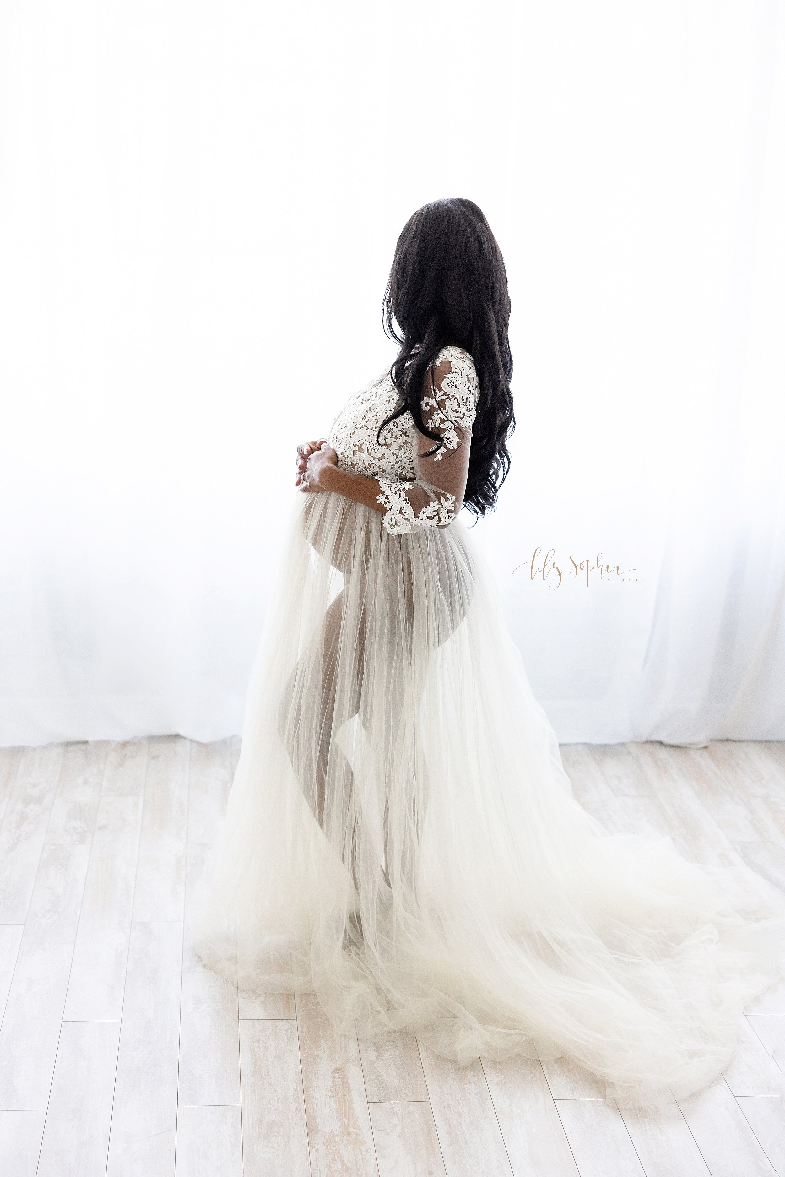  Maternity profile portrait of an African-American mother as she stands looking out a window streaming natural light wearing a lace bodice sheer skirted full-length gown with her hands resting on the top of her belly taken by Lily Sophia Photography 
