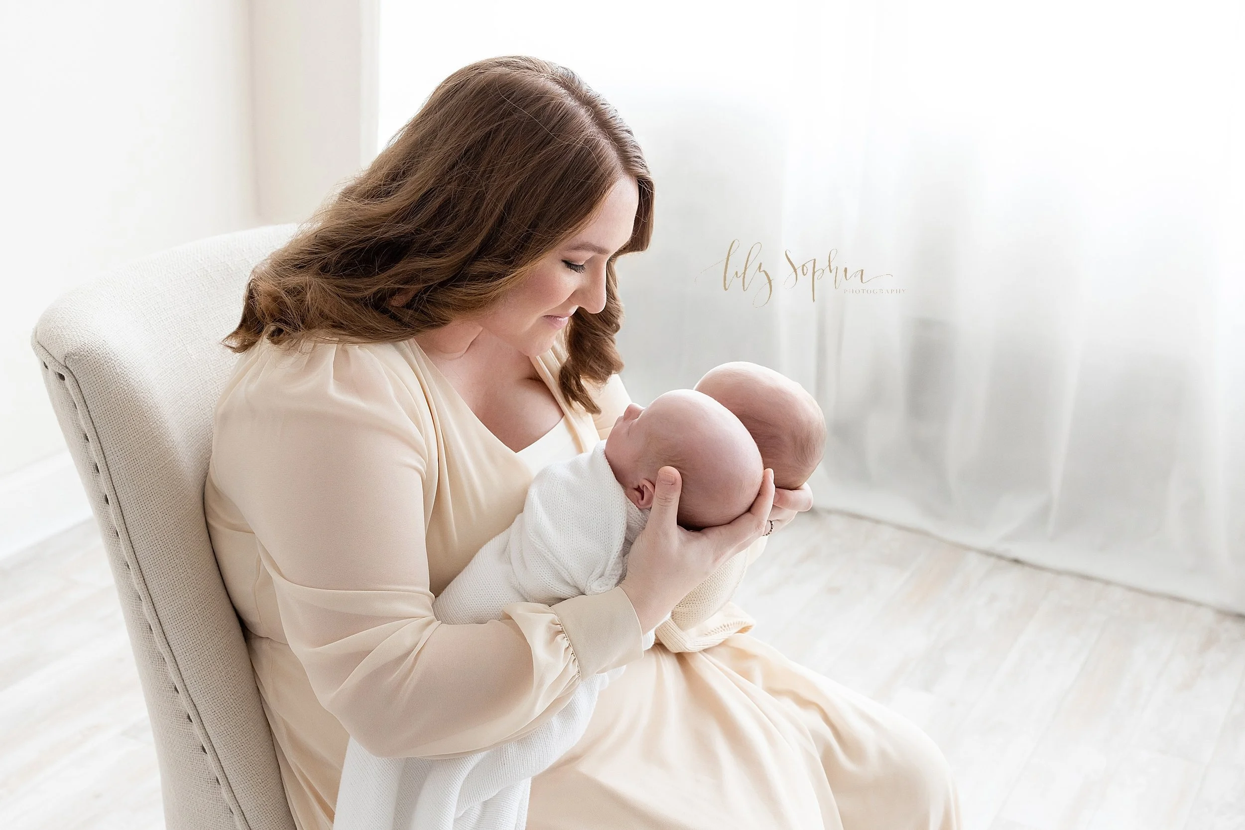  Newborn photograph of a mother sitting in an upholstered chair in front of a window streaming natural light in a studio as she holds her twin infant sons, one in each hand and admires them taken in Ponce City Market in Atlanta. 