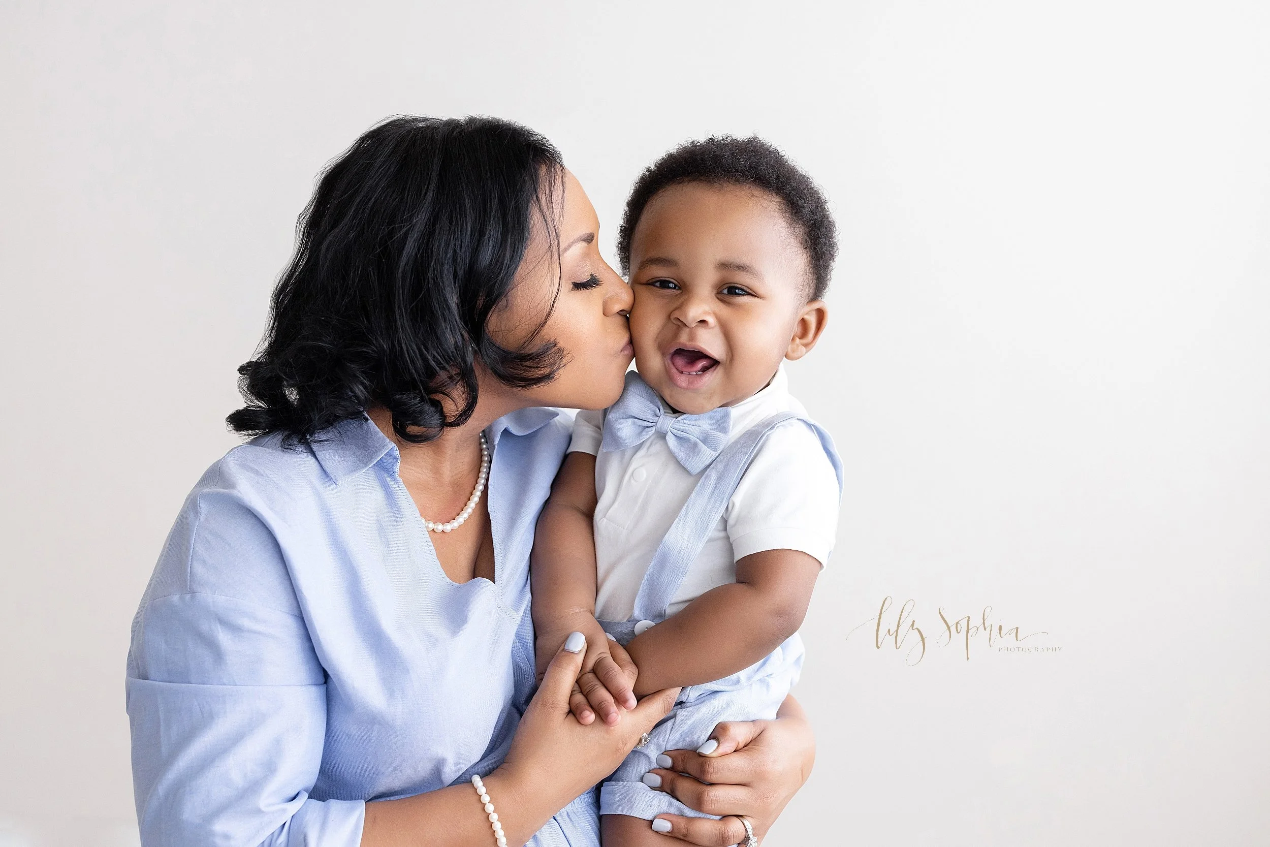  Family photograph of an African-American mother who is holding her one year old son in her arms as she kisses him on the cheek taken in near Kirkwood in Atlanta in a studio utilizing natural lighting. 