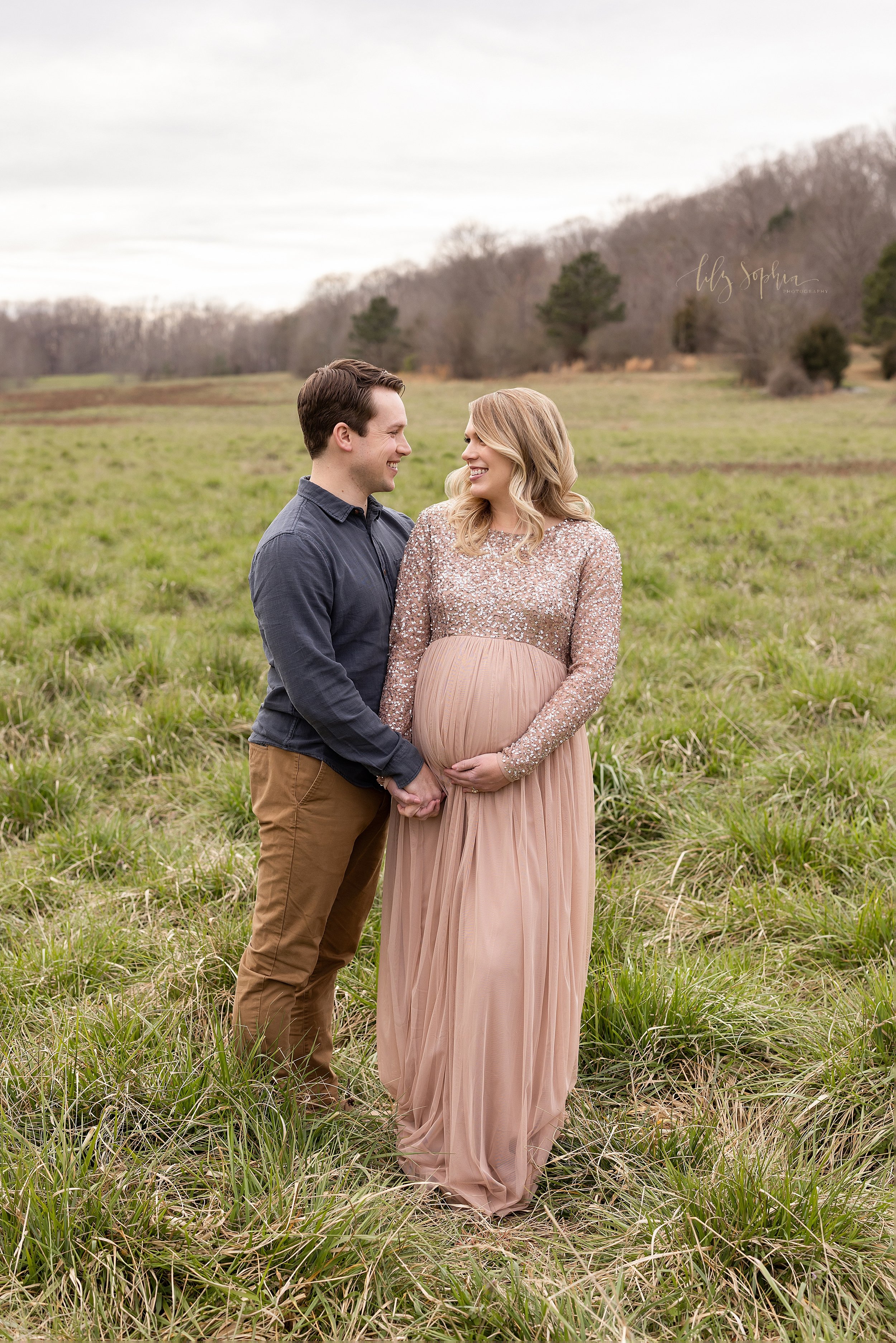  Maternity photo session during winter in a field near Atlanta of an expectant couple as the husband stands next to his wife holding her left hand with his right hand and his wife holds the base of her belly with her right hand as they smile at one a