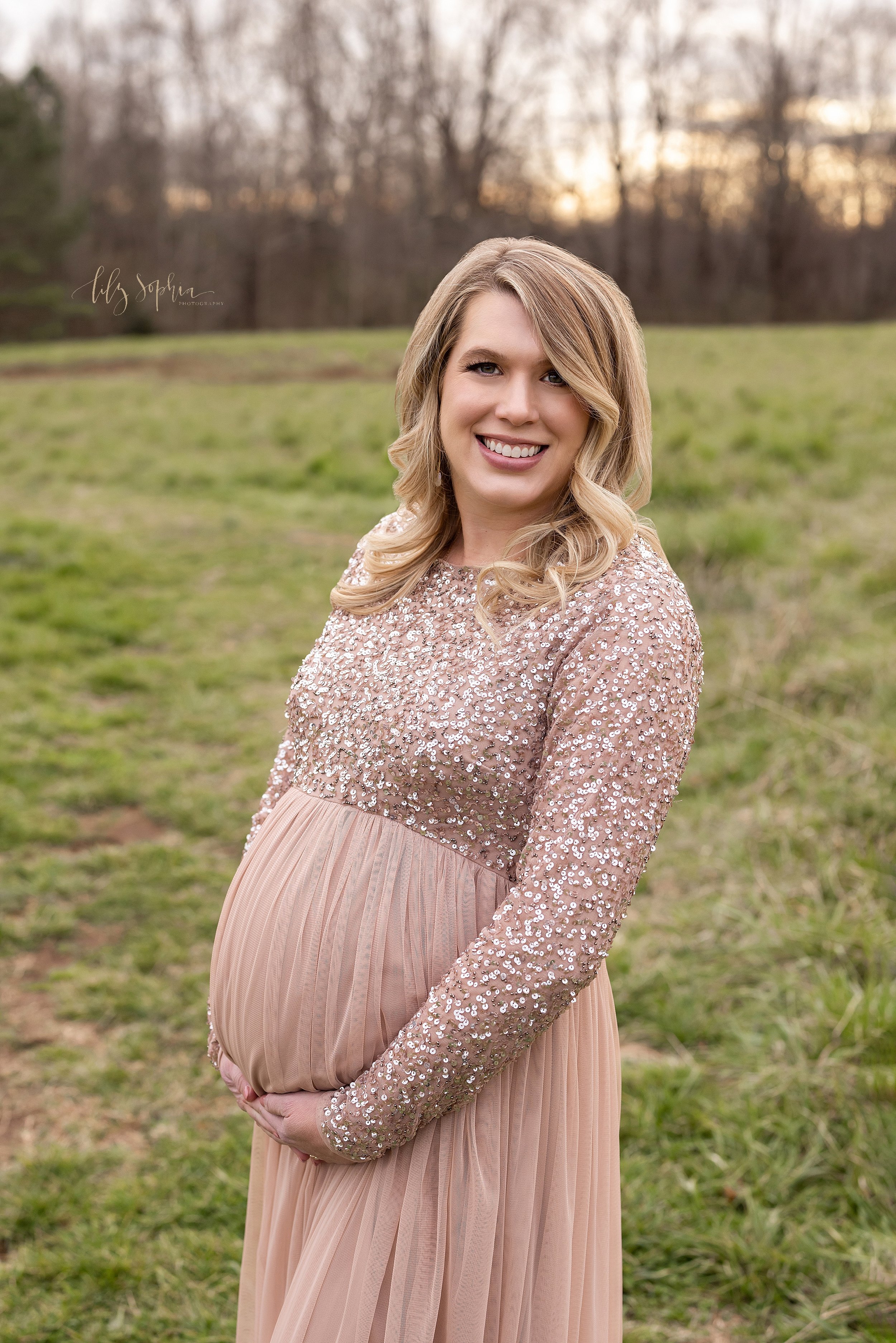  Maternity portrait of a glowing expectant mother wearing a long-sleeved, full-length gown with a sequined bodice as she holds the base of her belly and stands in a field near Atlanta, Georgia at sunset. 