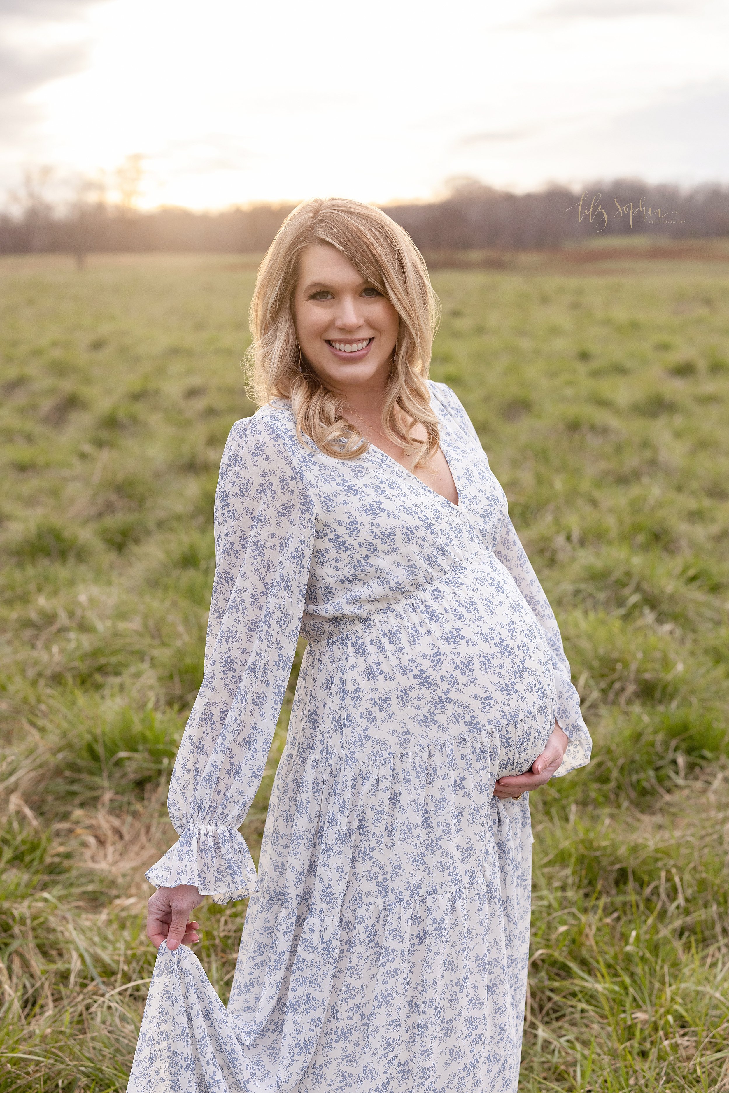  Maternity portrait of a pregnant mother standing with the sun setting behind her holding the base of her belly with her left hand and the hem of her cotton long sleeved printed gown with her right hand in an Atlanta field. 