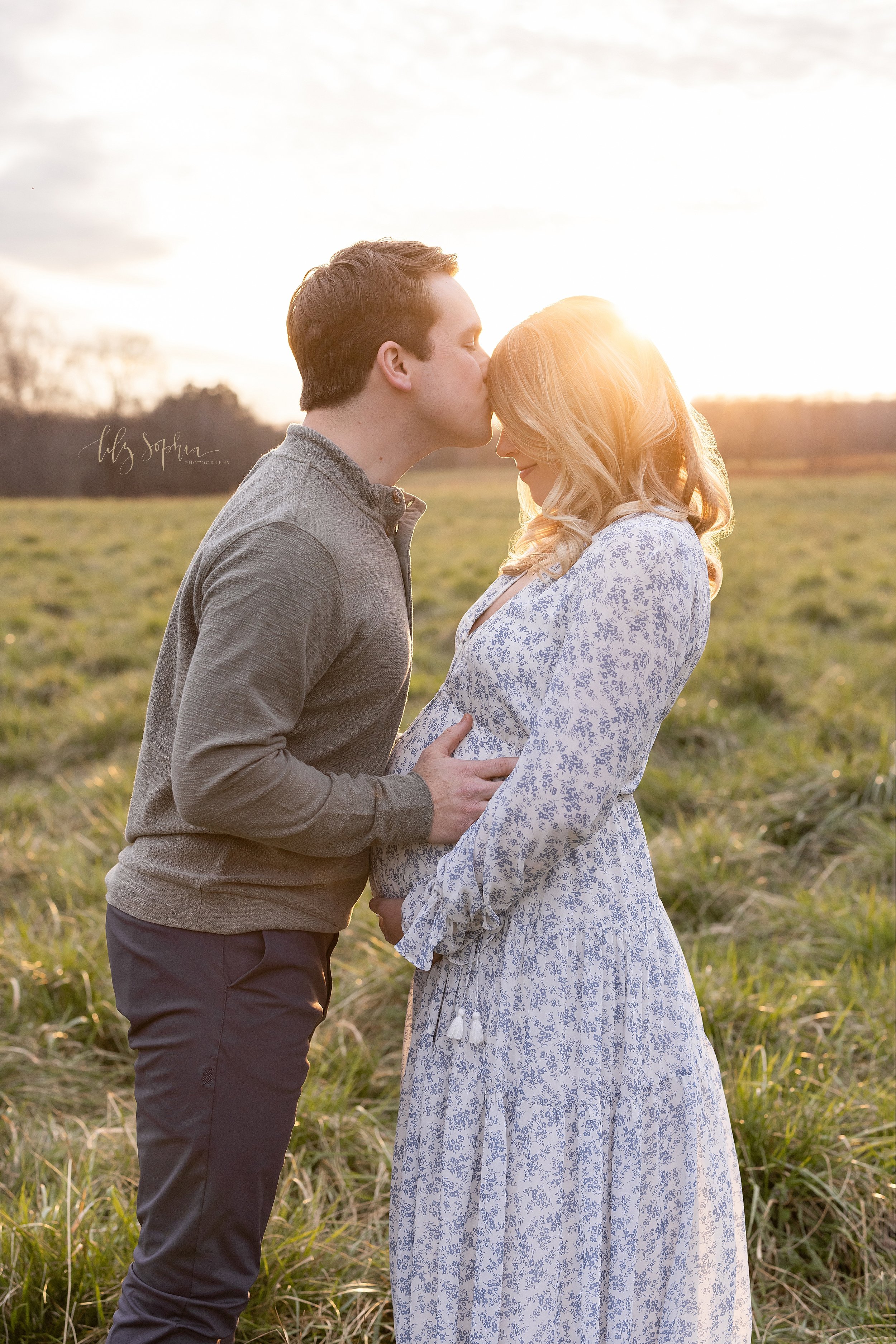  Maternity portrait of the love between husband and wife as the husband faces his wife and kisses her on her forehead while placing his hands on his child in utero as the couple stands with the sun setting behind them in a field near Atlanta, Georgia