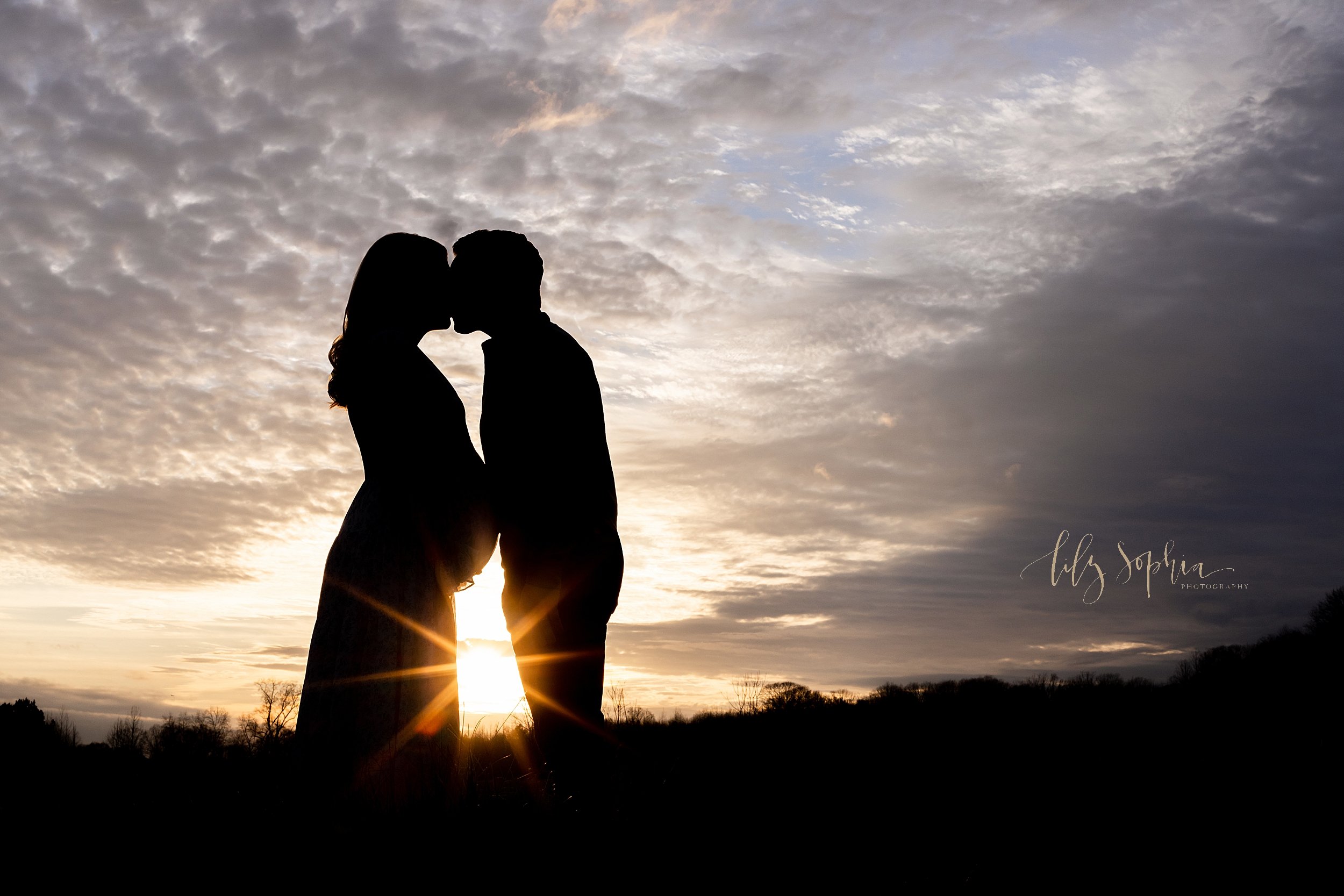  Silhouette maternity portrait of an expectant couple as they stand at sunset in an Atlanta field facing one another and kiss each other. 