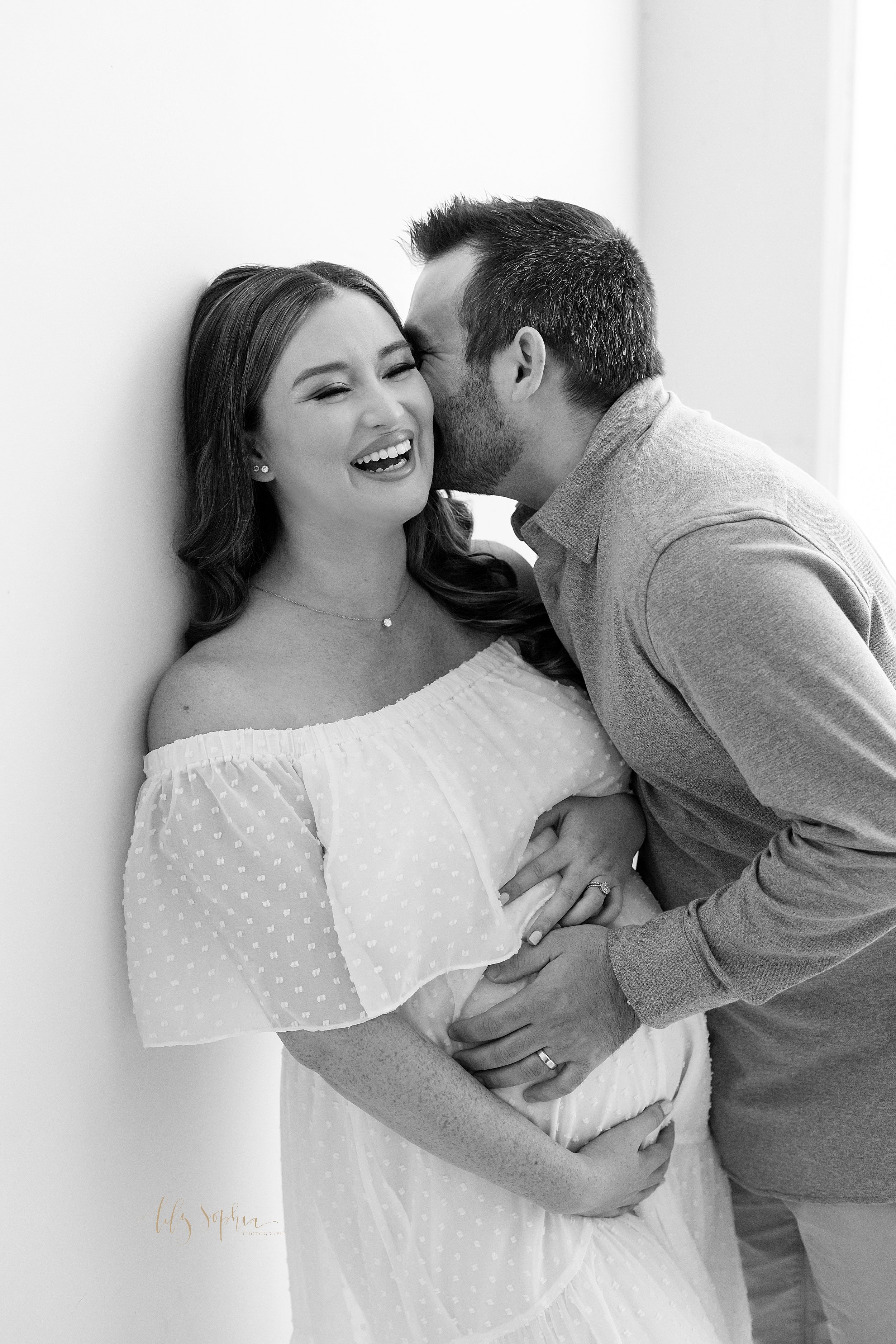 Maternity photo of a laughing pregnant mother as her husband touches her belly with his hands and whispers in her ear taken in a studio near Poncey Highlands in Atlanta. 