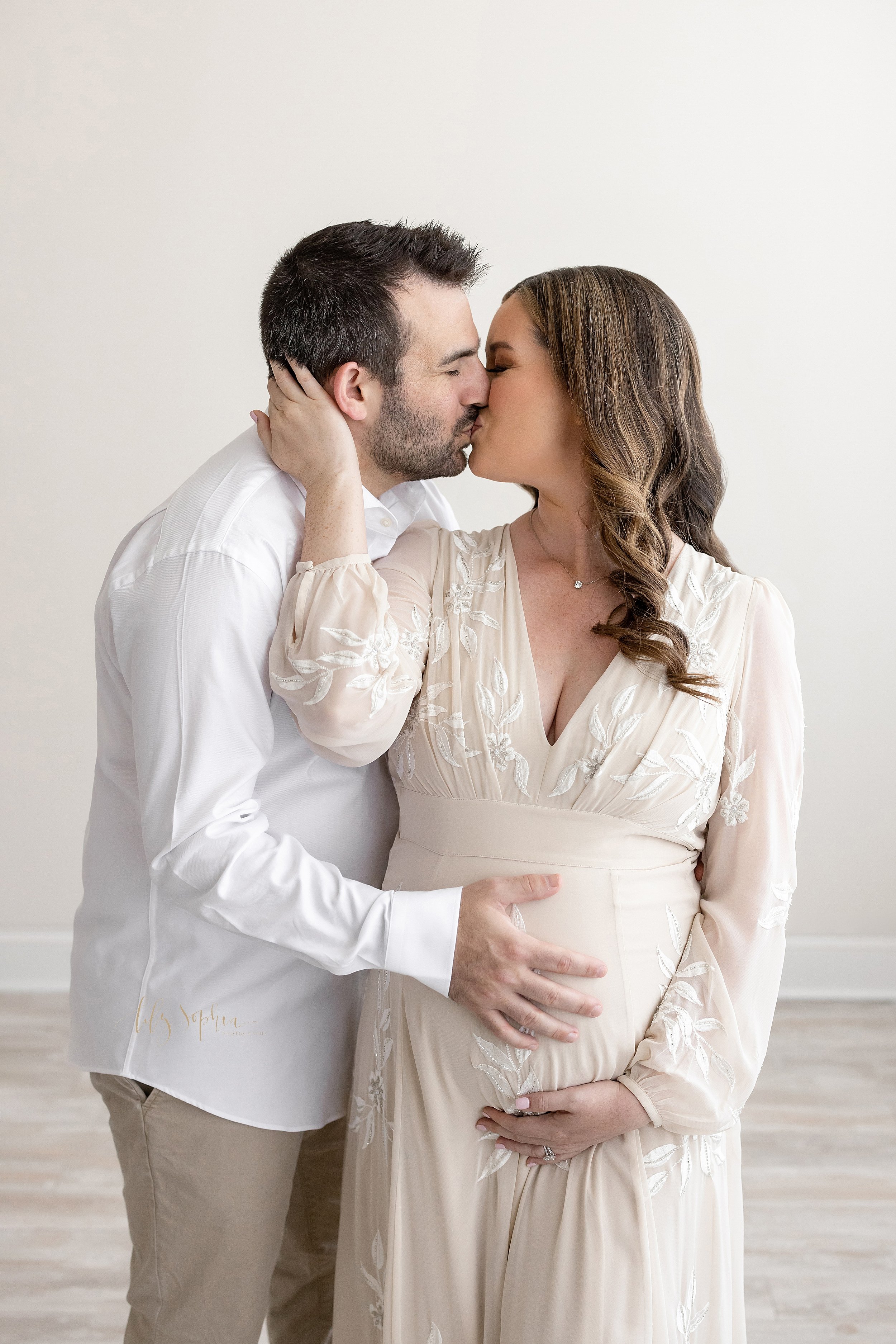  Maternity portrait of the love between husband and wife as they stand in a studio with the wife turning her head over her right shoulder and placing her right hand on her husband’s neck to kiss him while he places his right hand on their child in ut