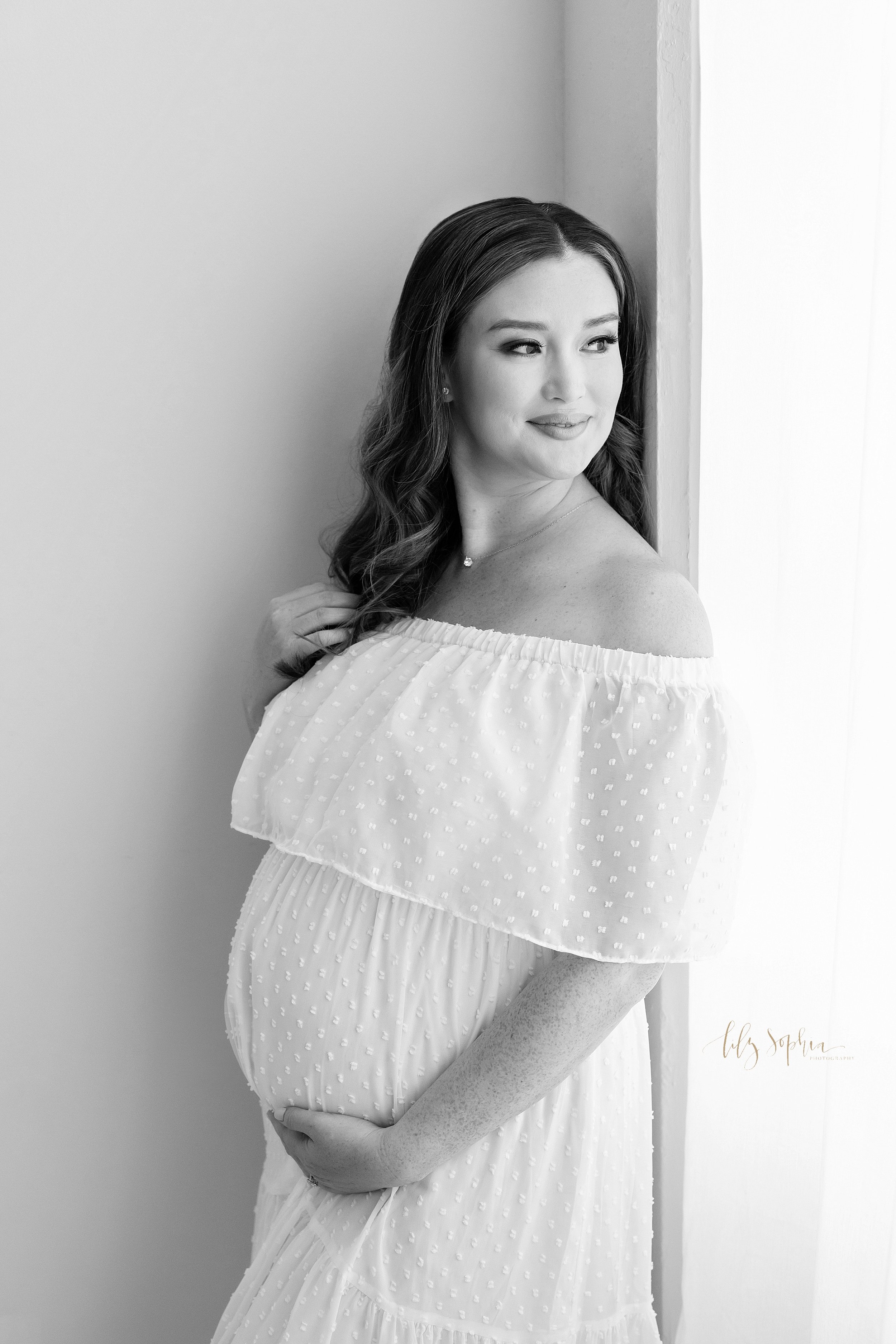 Maternity portrait in black and white of an expectant mother as she stands with her right hand touching her hair and her left hand holding her pregnant belly while she looks over her left shoulder out a window in a studio near Decatur that uses natu