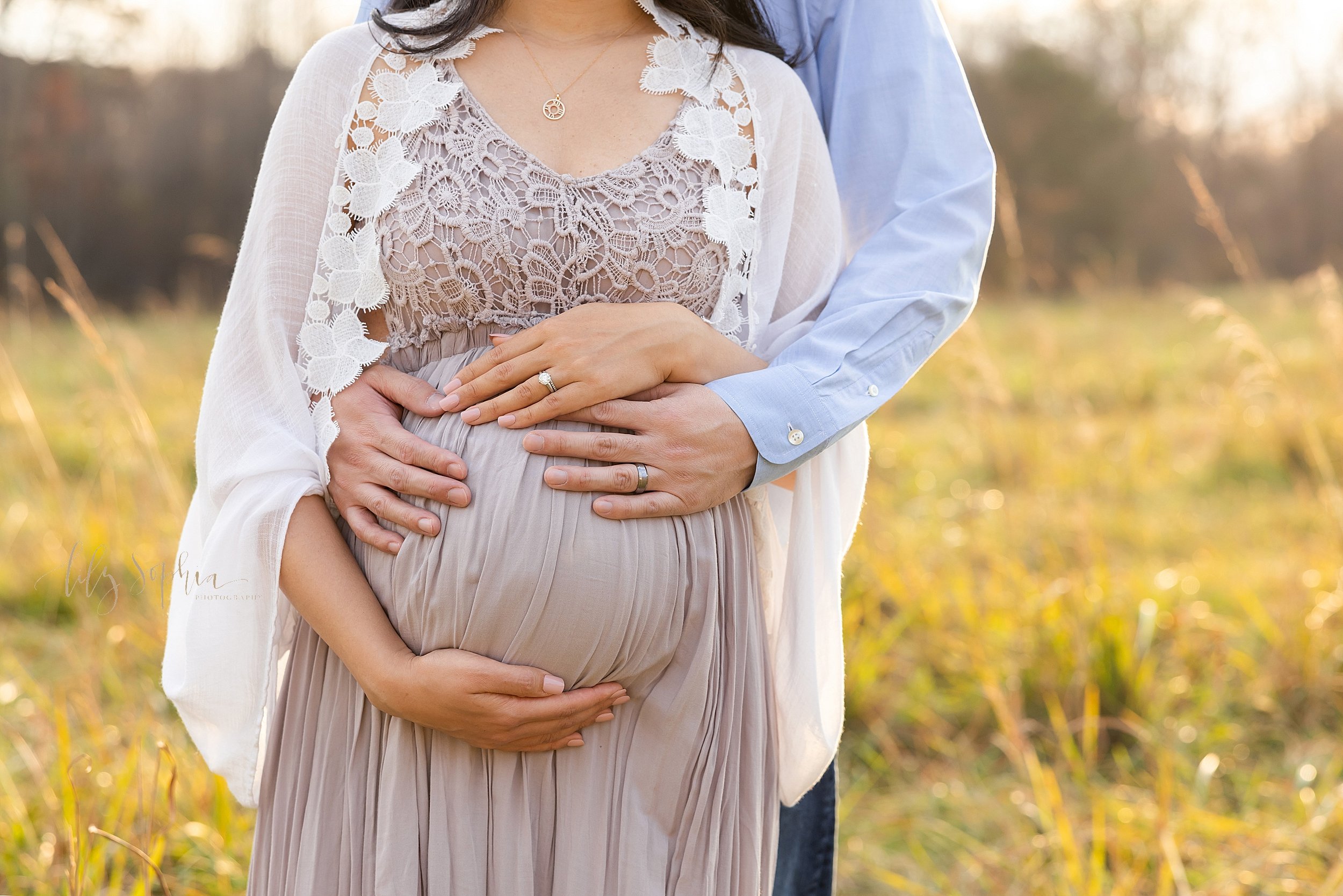 Close-up maternity photo of a husband standing behind his wife embracing his child in utero as his wife frames her belly with her hands taken in autumn in an Atlanta field at sunset. 
