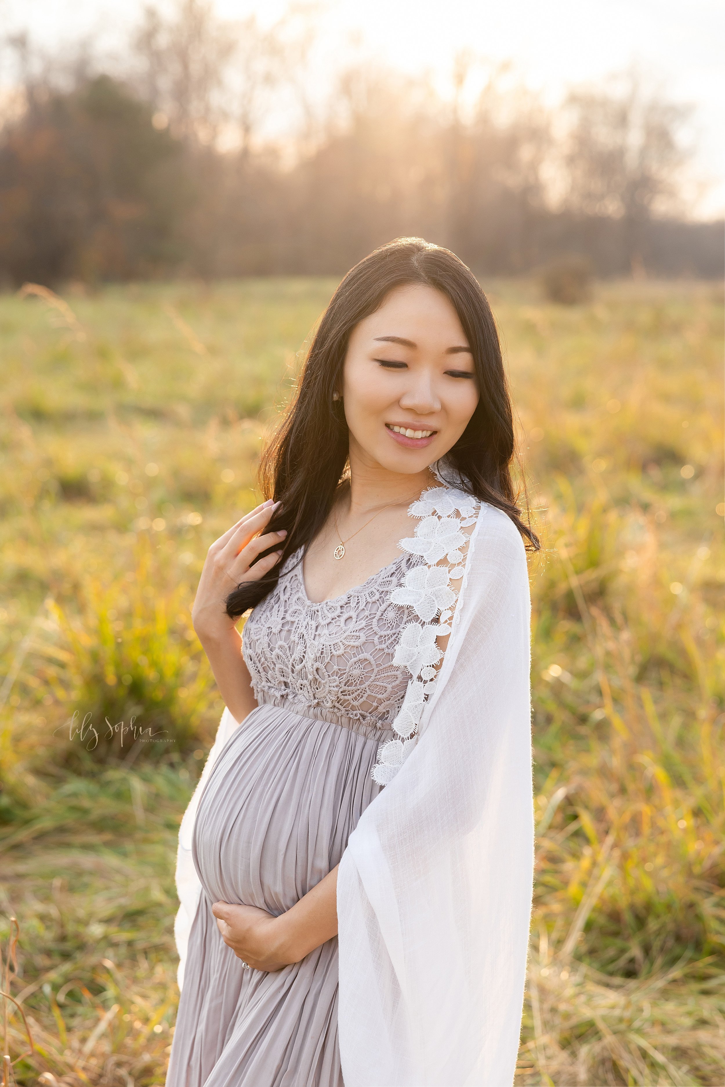  Maternity portrait of a pregnant Asian mother as she stands in a field near Atlanta at sunset with her left hand holding the base of her belly and her right hand touches her hair while she looks over her left shoulder. 