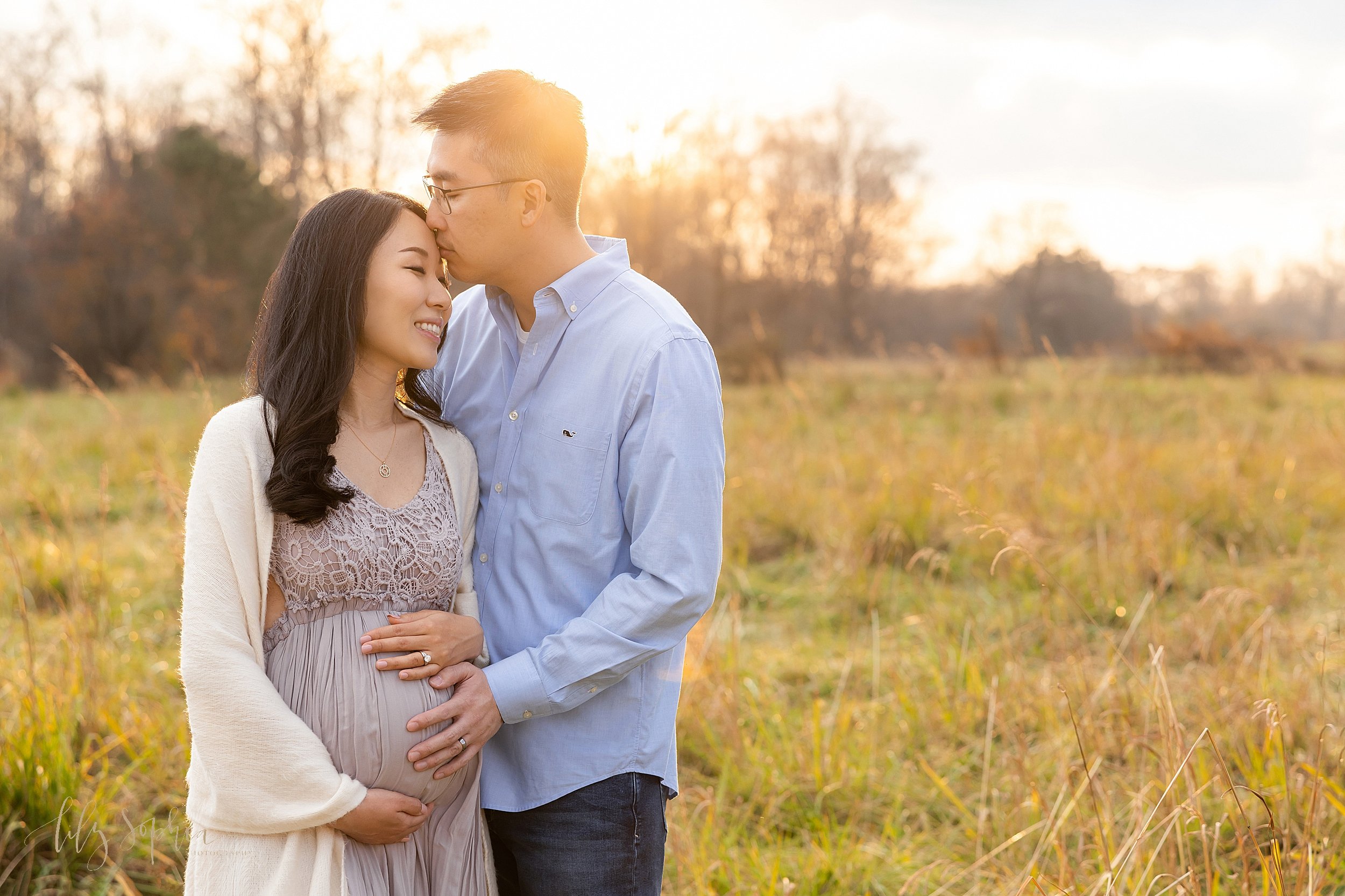  Maternity photo taken in an Atlanta field of an Asian couple with the husband standing to the right of his wife as he places his left hand on his wife’s belly and kisses her on the forehead taken as the sun sets during autumn. 