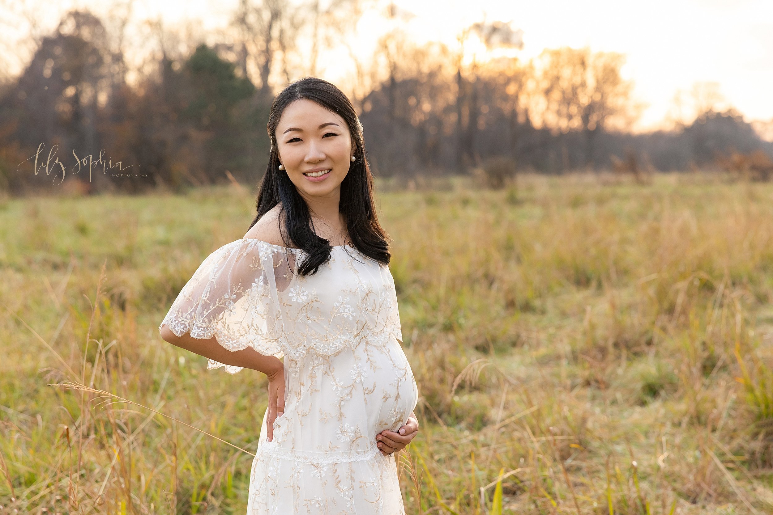  Maternity portrait of an Asian mother as she stands at sunset wearing an off-the shoulder lacy gown with her right hand on her hip and her left hand holding the base of her belly during the fall season taken in an Atlanta field. 