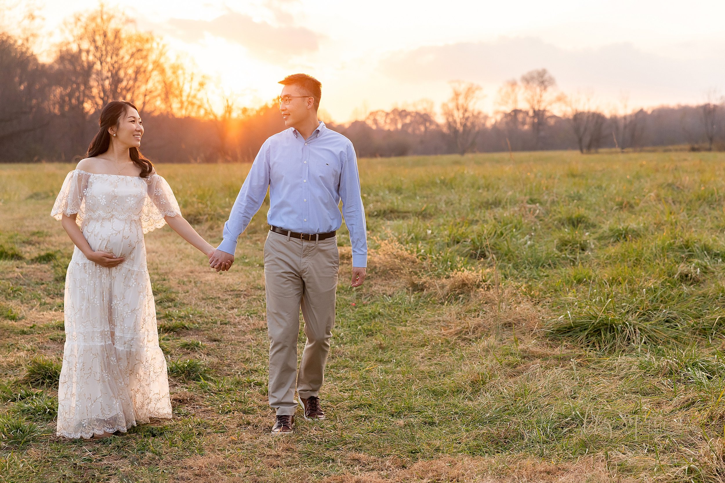  Maternity photo of an expectant Asian couple walking hand in hand through a field with the sun setting between them during the fall season near Atlanta, Georgia. 