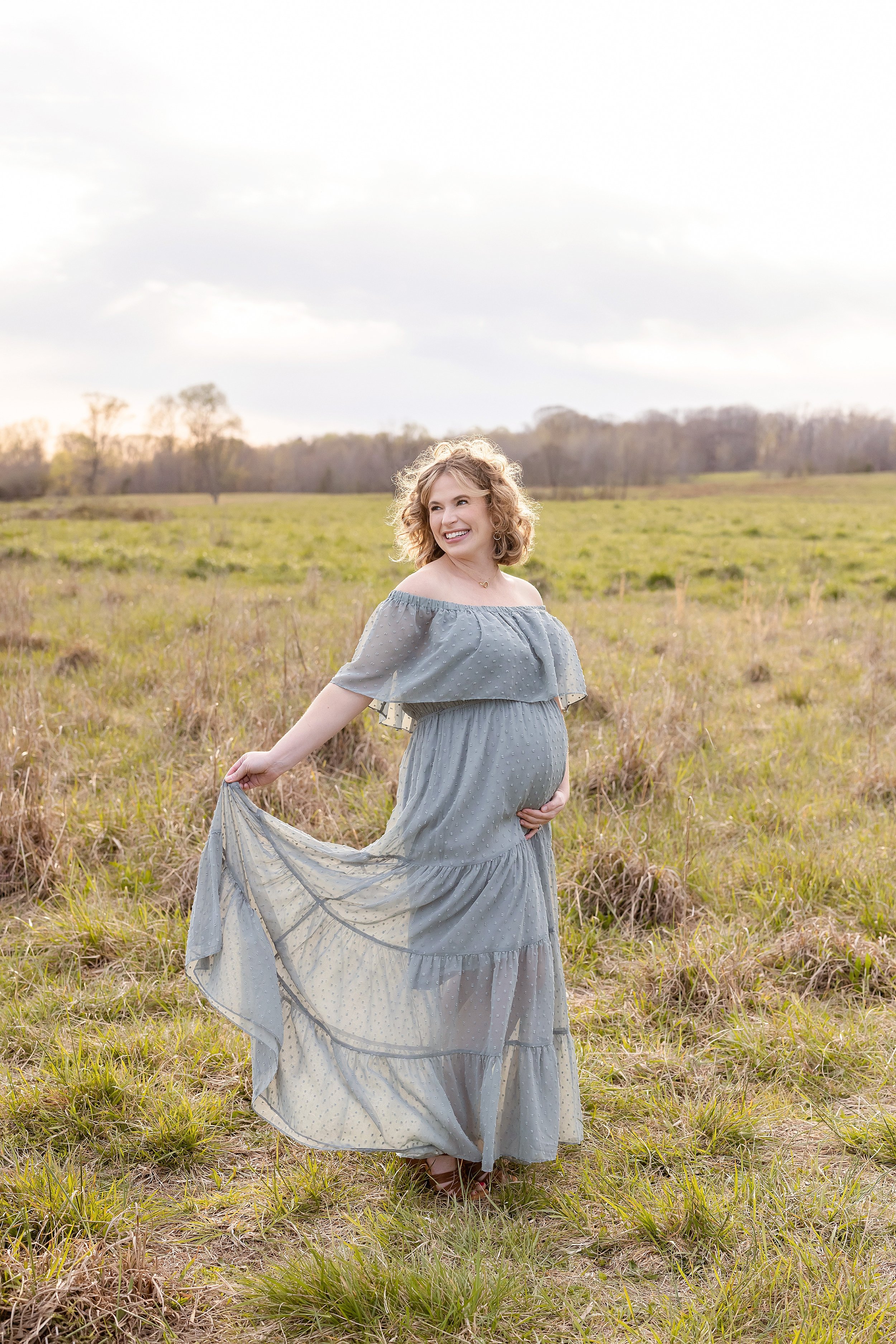  Maternity portrait of a radiant expectant mother wearing an off-the-shoulder wide-bodice flowing gown as she stands at sunset in a field during autumn holding the skirt of her gown in her right and the base of her belly with her left hand as she smi