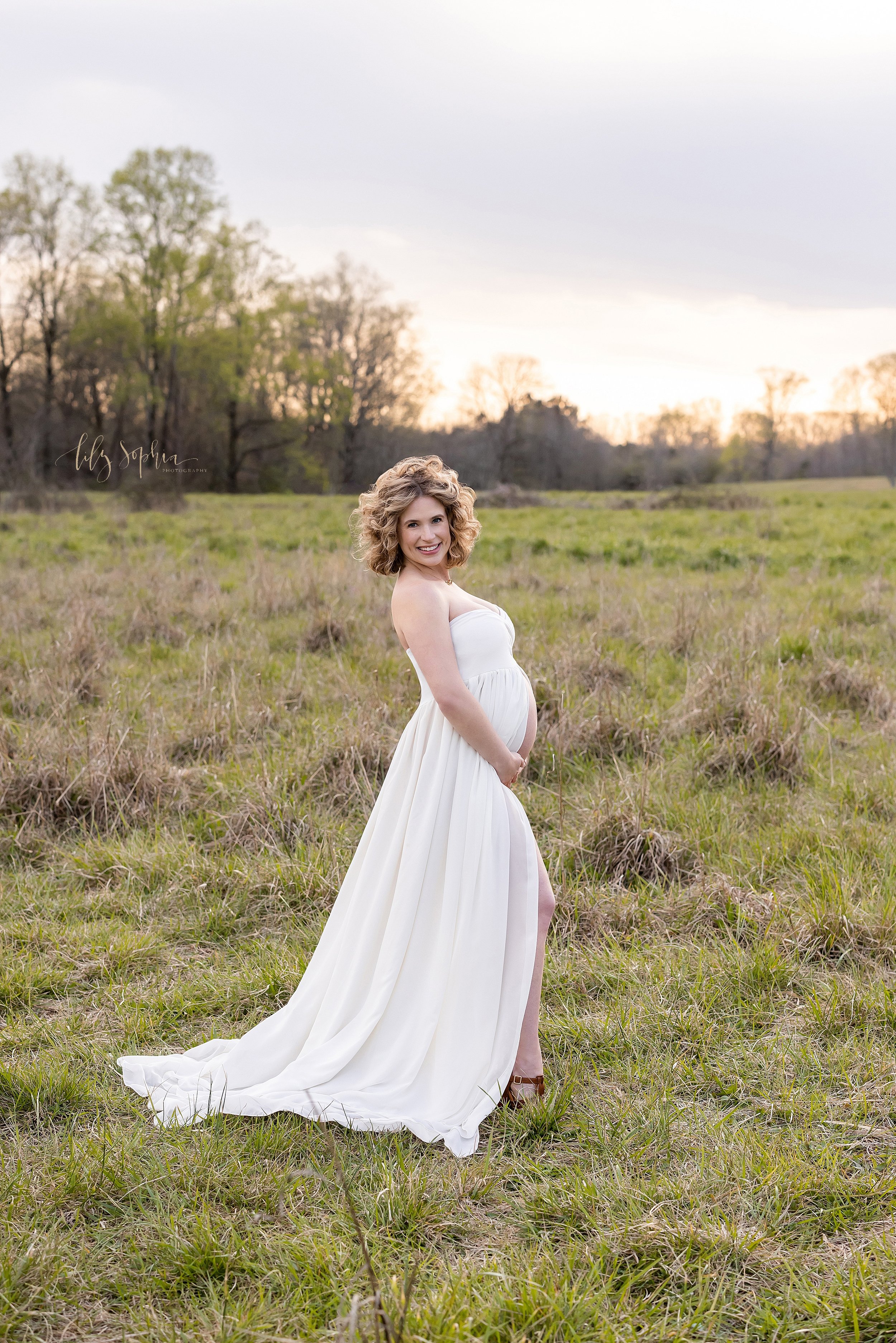  Maternity portrait of a pregnant mother wearing a split front full-length gown as she stands during winter in a field at sunset with her hands beneath her belly and bears her belly taken near Atlanta. 