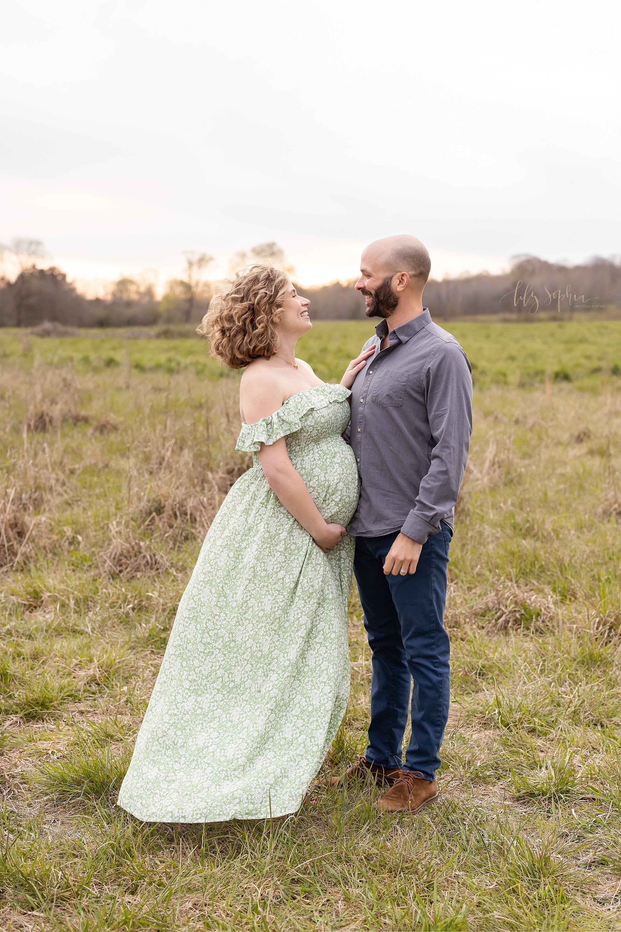  Maternity photograph of a mother and father as they stand facing one another in a field with the mother placing her right hand on her husband’s chest and her left hand holding the base of her belly as the couple smile at one another taken during the