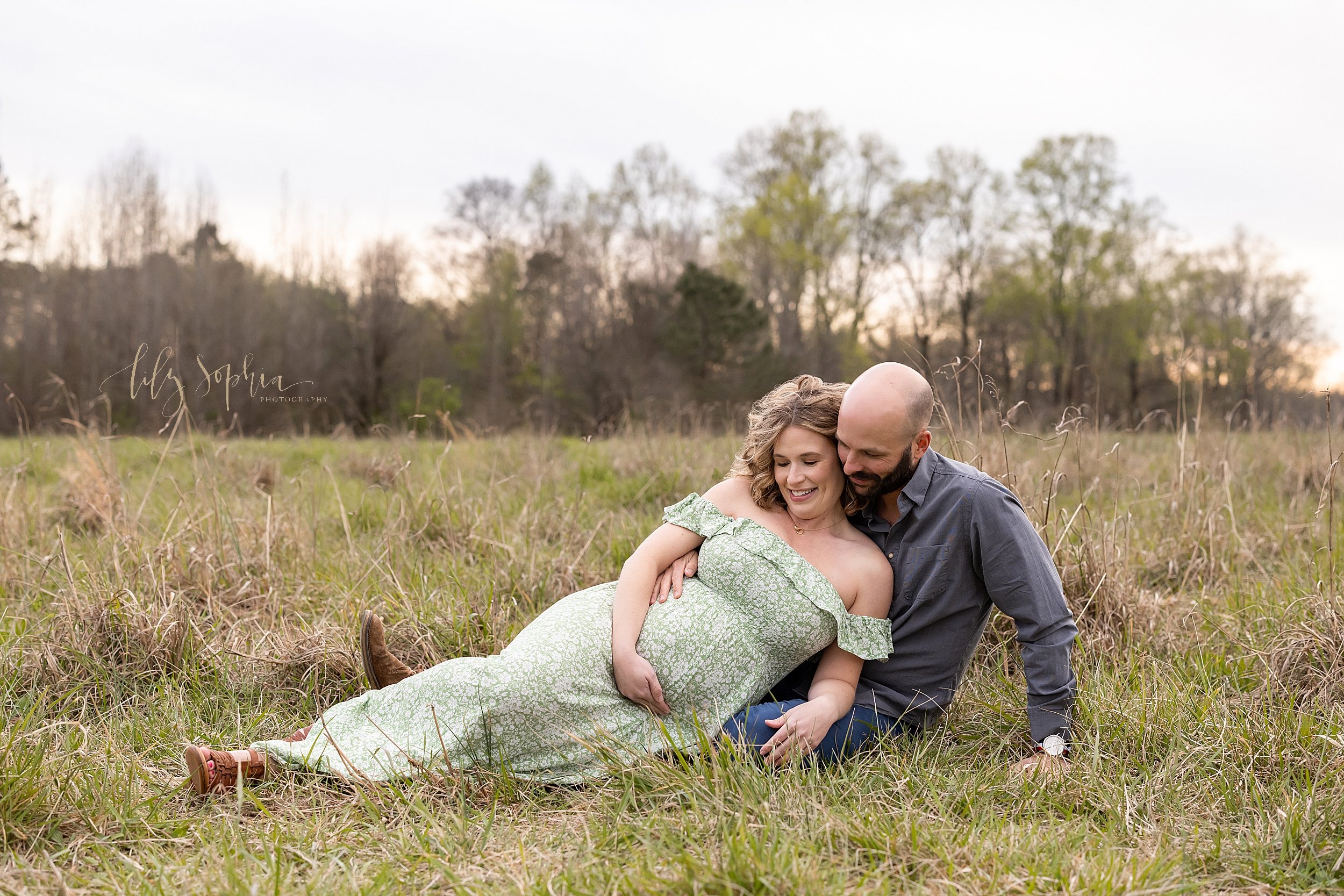  Maternity photo taken in a field near Atlanta, Georgia during winter of an expectant couple lounging on the grassy field with the mother wearing an off the shoulder printed dress as she leans against her husband while holding the base of her belly a