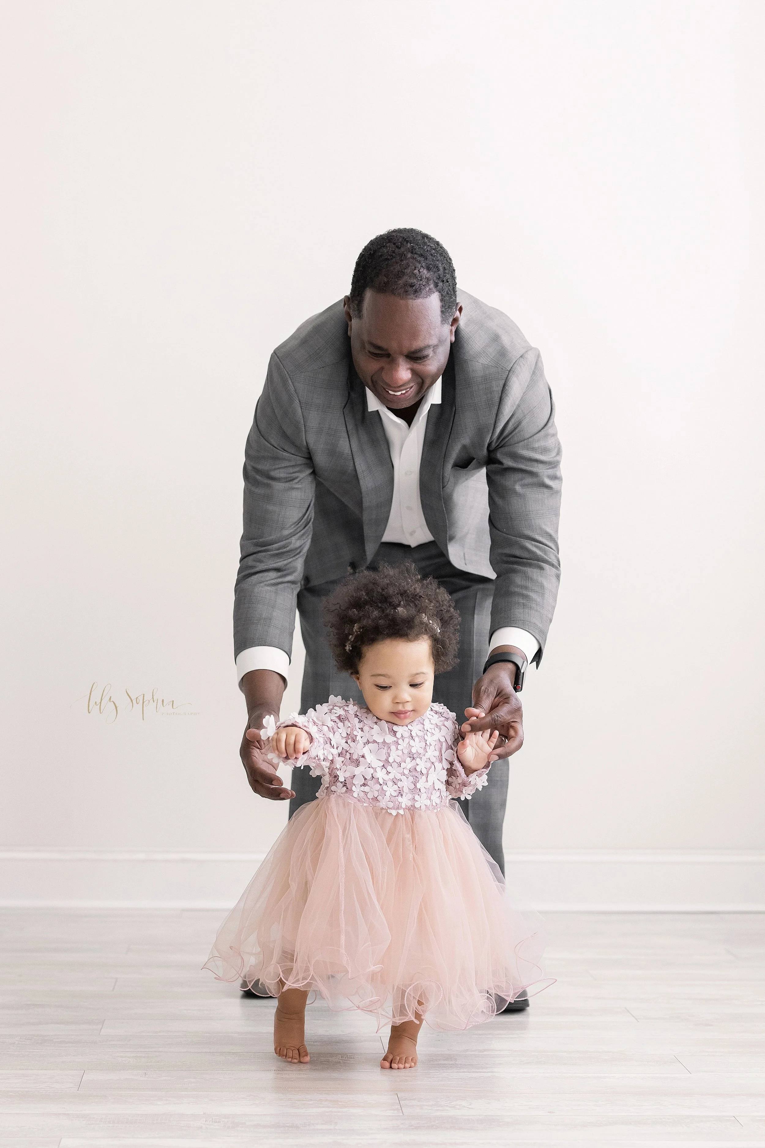  First birthday photograph of an African-American  one year old little girl as she holds her father’s fingers while walking in front of him in a natural light studio near Morningside in Atlanta, Georgia. 