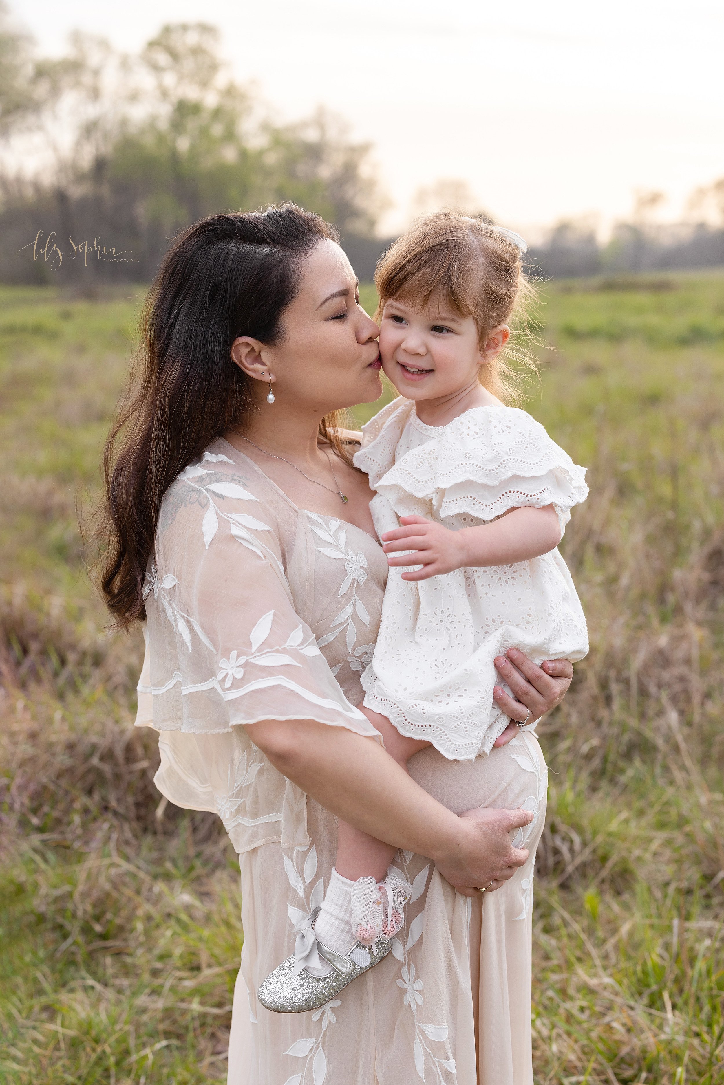  Maternity mother-daughter portrait as the expectant mother holds her young daughter in her arms resting on her belly as she kisses her daughter on her cheek as she stands in an Atlanta field at sunset. 