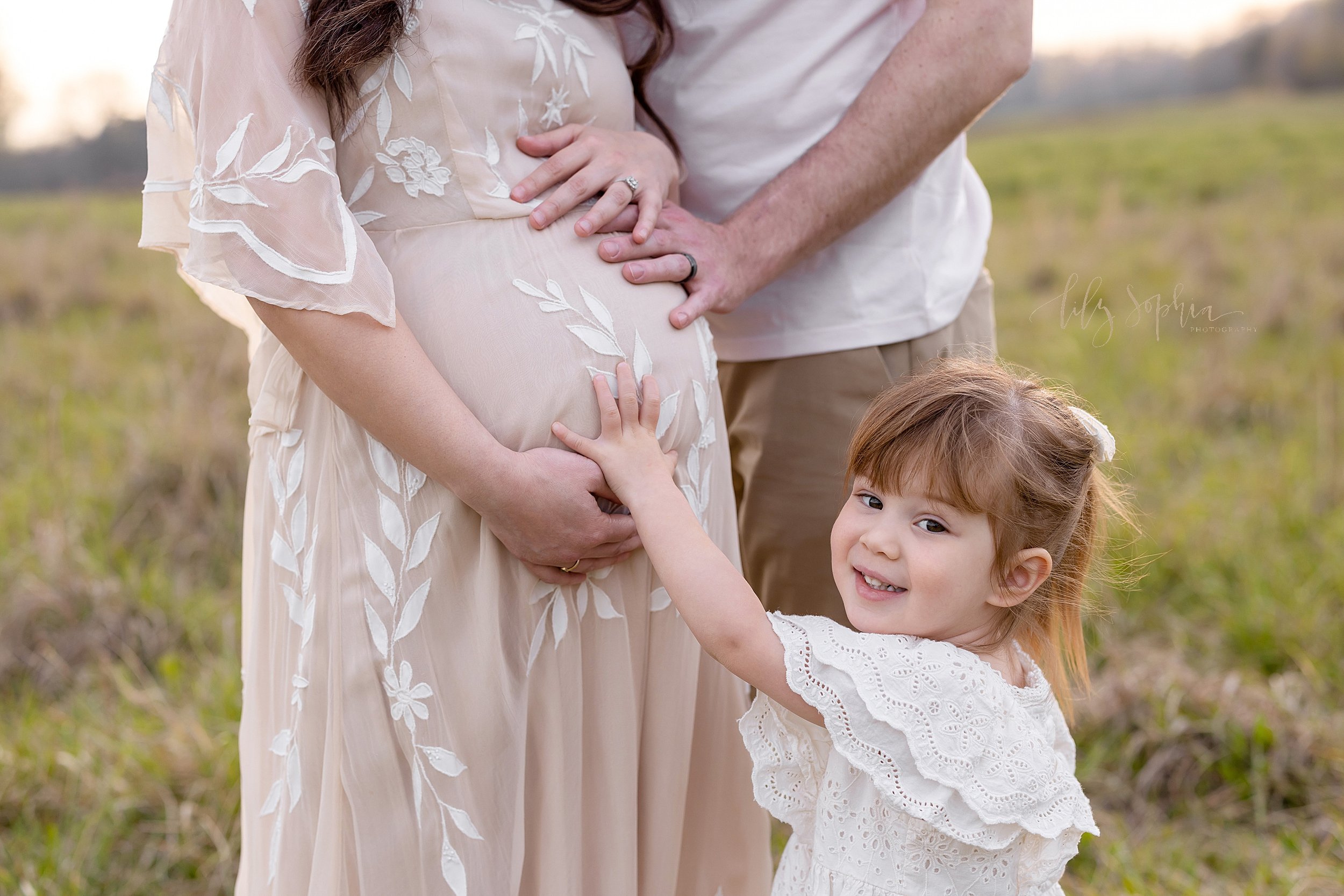  Maternity family photograph of an expectant mother standing in a field with her hands framing her belly  and her husband standing to her left side with his hand on their child in utero while their young daughter stands in front of her mother as she 