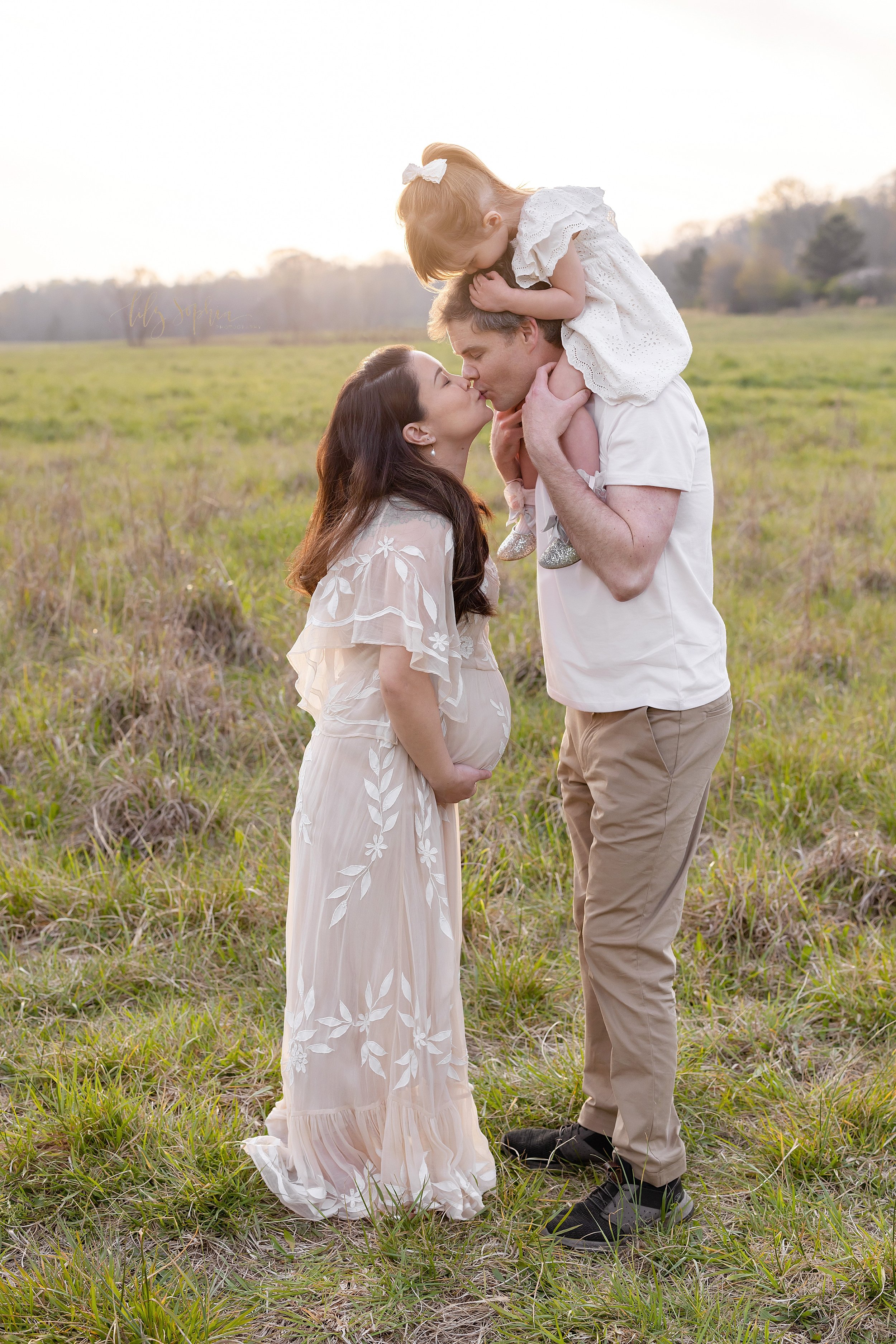  Family maternity photo with a pregnant mother standing facing her husband who is holding their young daughter on his shoulders as mom kisses her husband while holding her belly and the little girl kisses her father’s head as the sun sets behind them