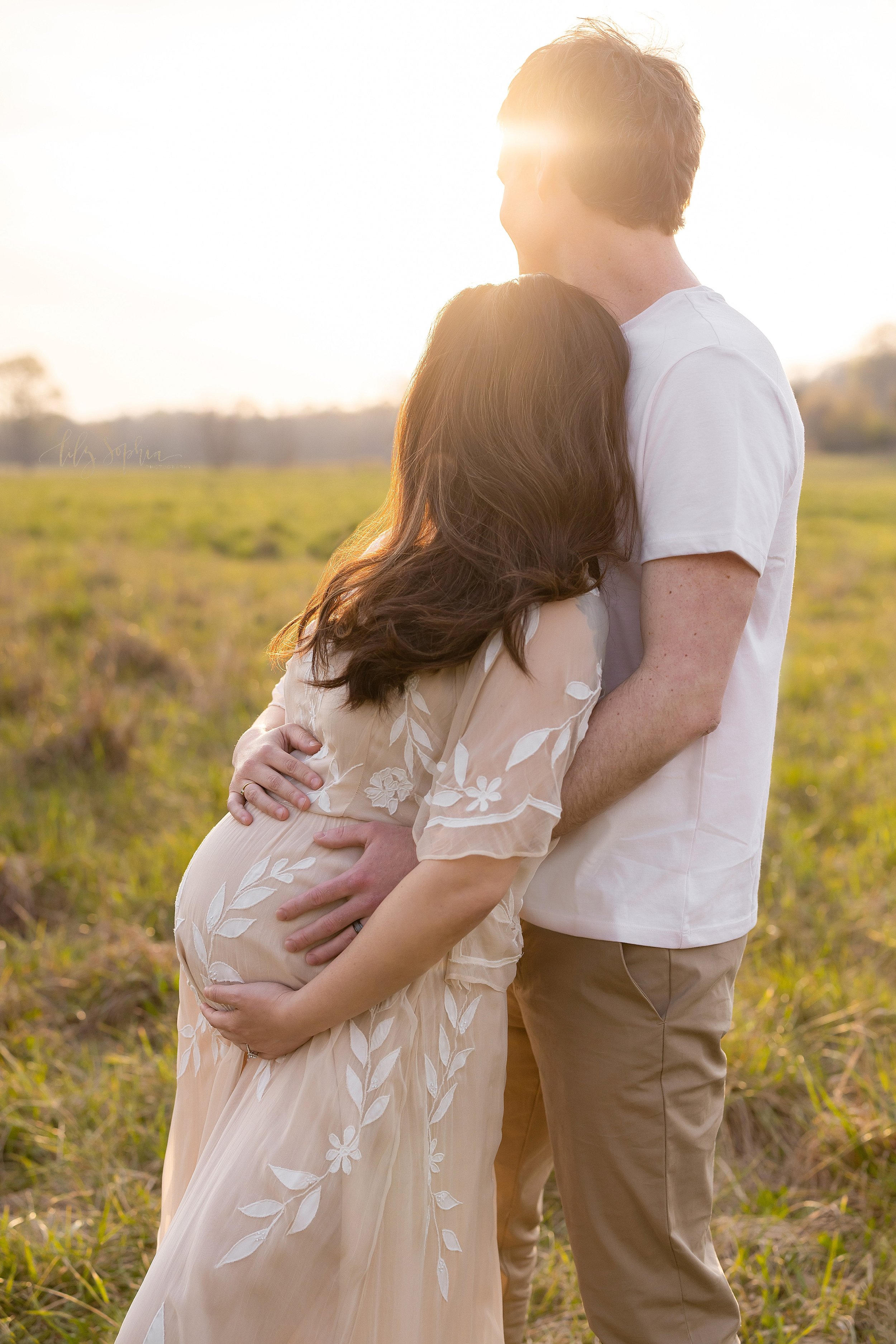  Maternity photo taken at sunset in a field near Atlanta of a pregnant mother framing her belly as she stands with her back against her husband’s chest as he wraps his hands around her waist to hold their child in utero. 