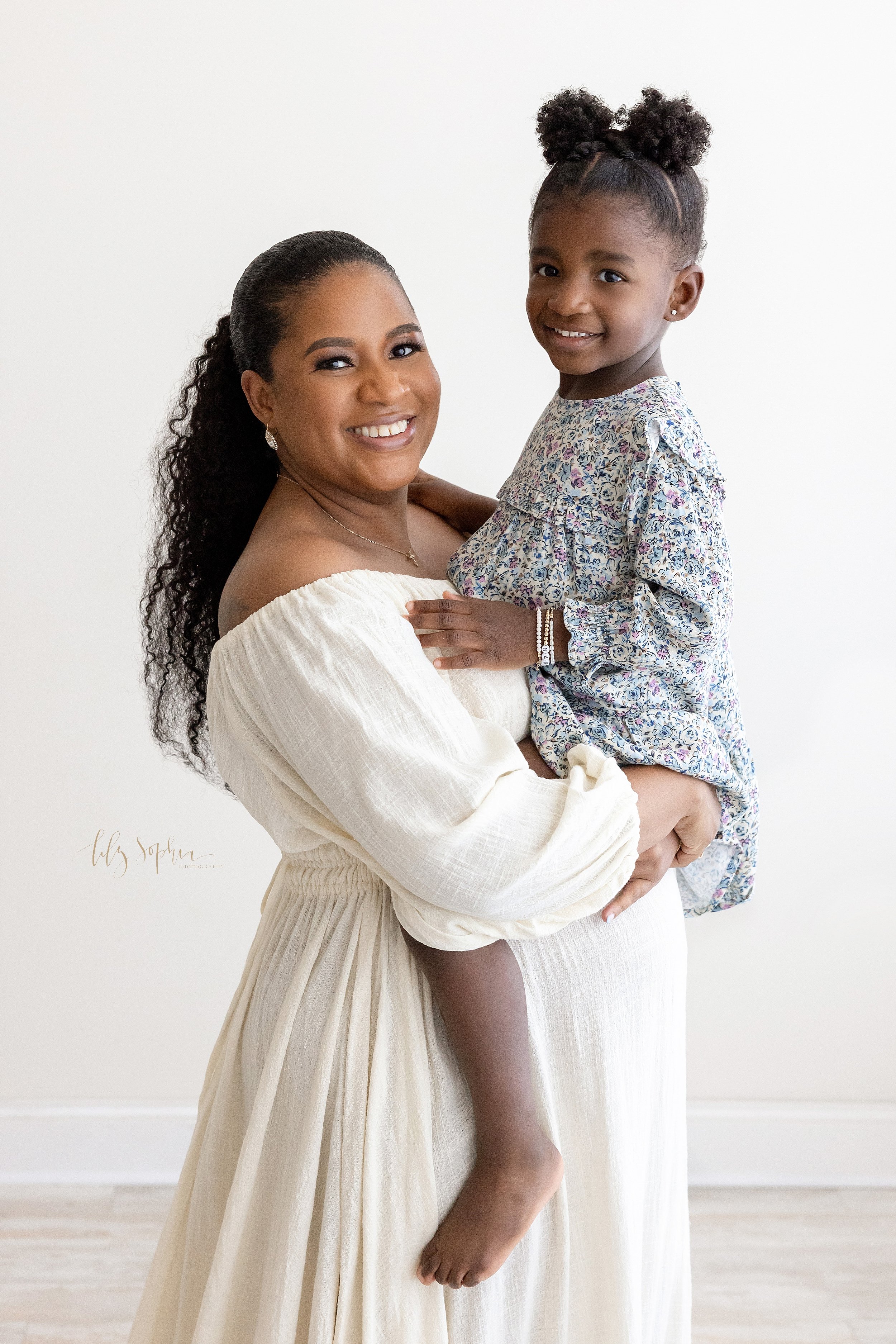  Maternity photo of an expectant African-American mom holding her young daughter as she stands in a natural light studio near Ansley Park in Atlanta, Georgia. 