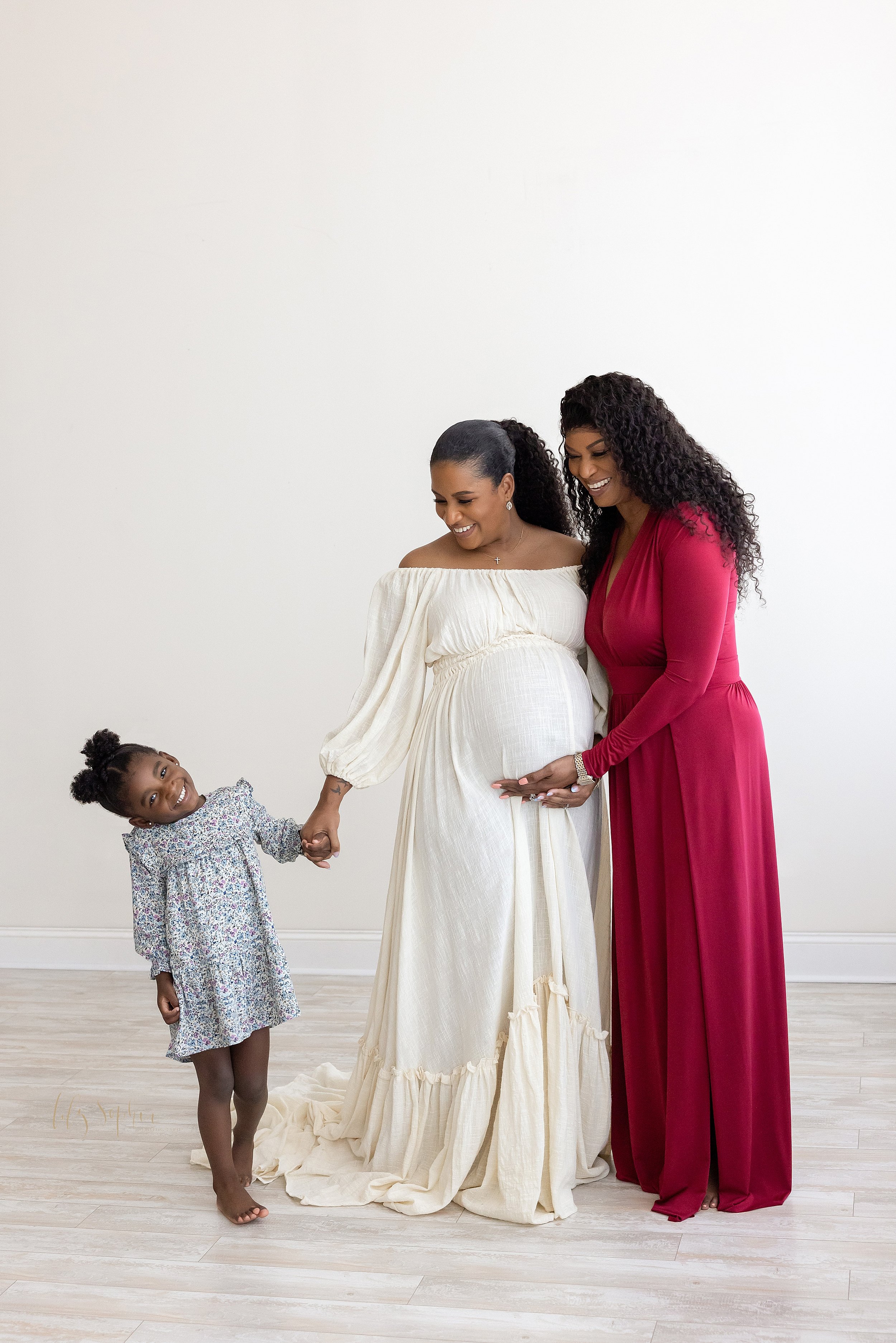  Three generation maternity photograph of an African-American expectant mother holding her young daughter’s hand with her right hand as her mother stands next to her on her left and the two of them frame her belly with their left hands taken in a nat