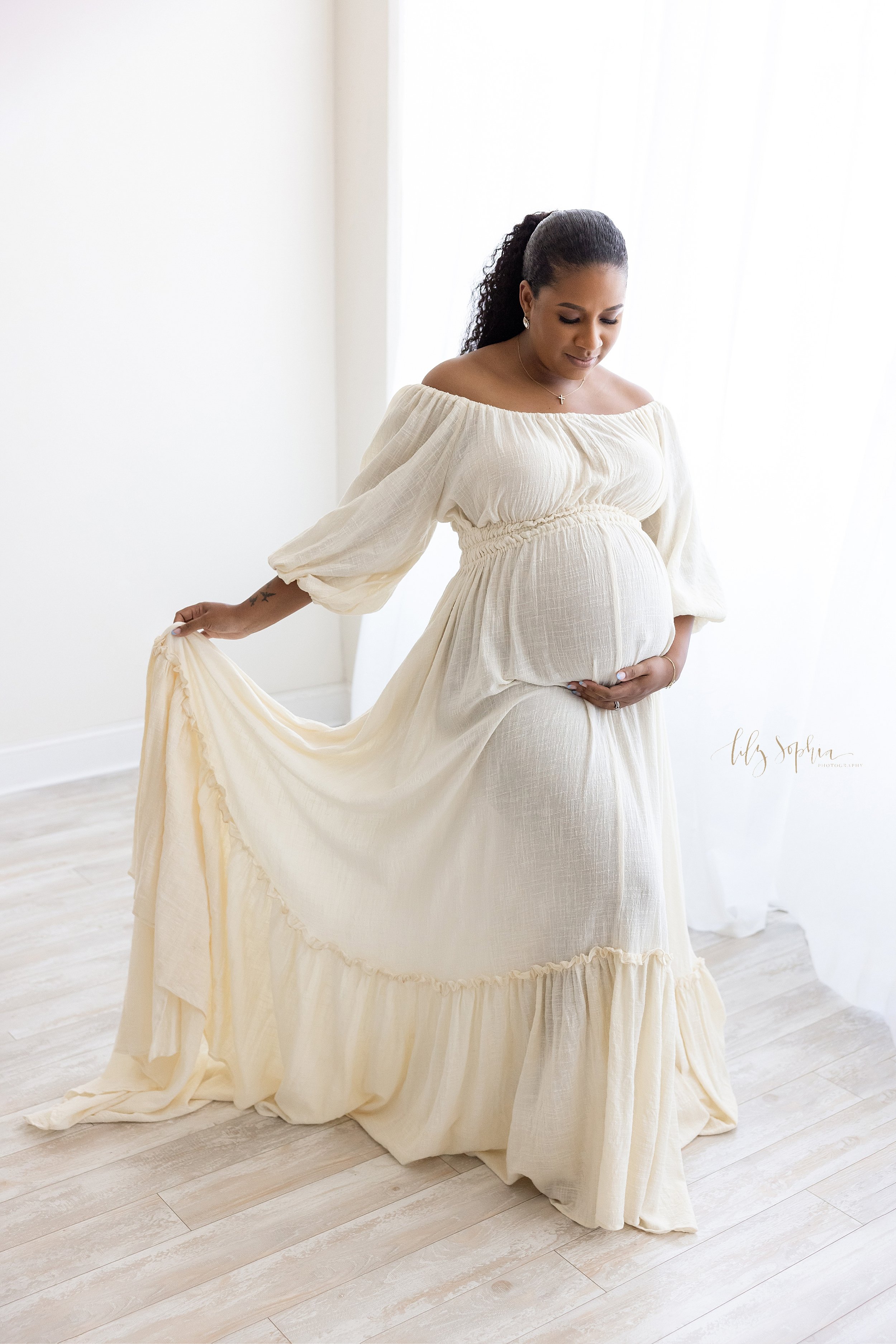  Maternity portrait of an African-American mother wearing an off the shoulder peasant sleeved linen full-length gown as she stands and fans the gown with her right hand while holding her belly with her left hand taken in front of a window streaming n