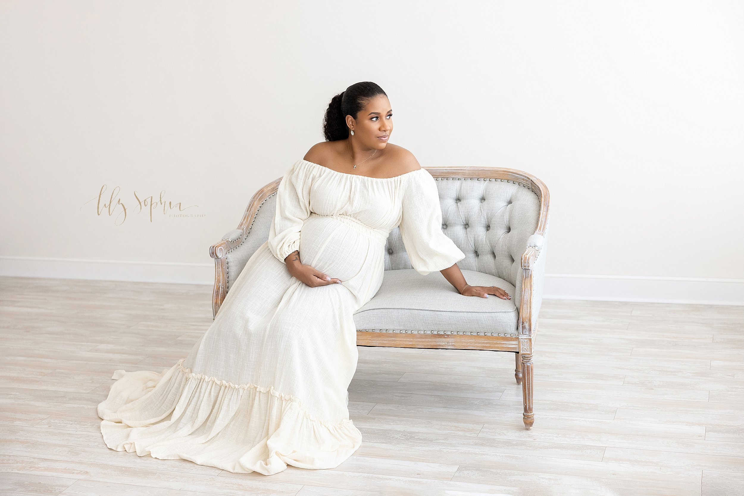  Maternity portrait of an expectant mother as she sits on a tufted backed love seat leaning on her left hand and holding her belly with her right hand as she looks out a natural light window in a studio near Brookhaven in Atlanta. 