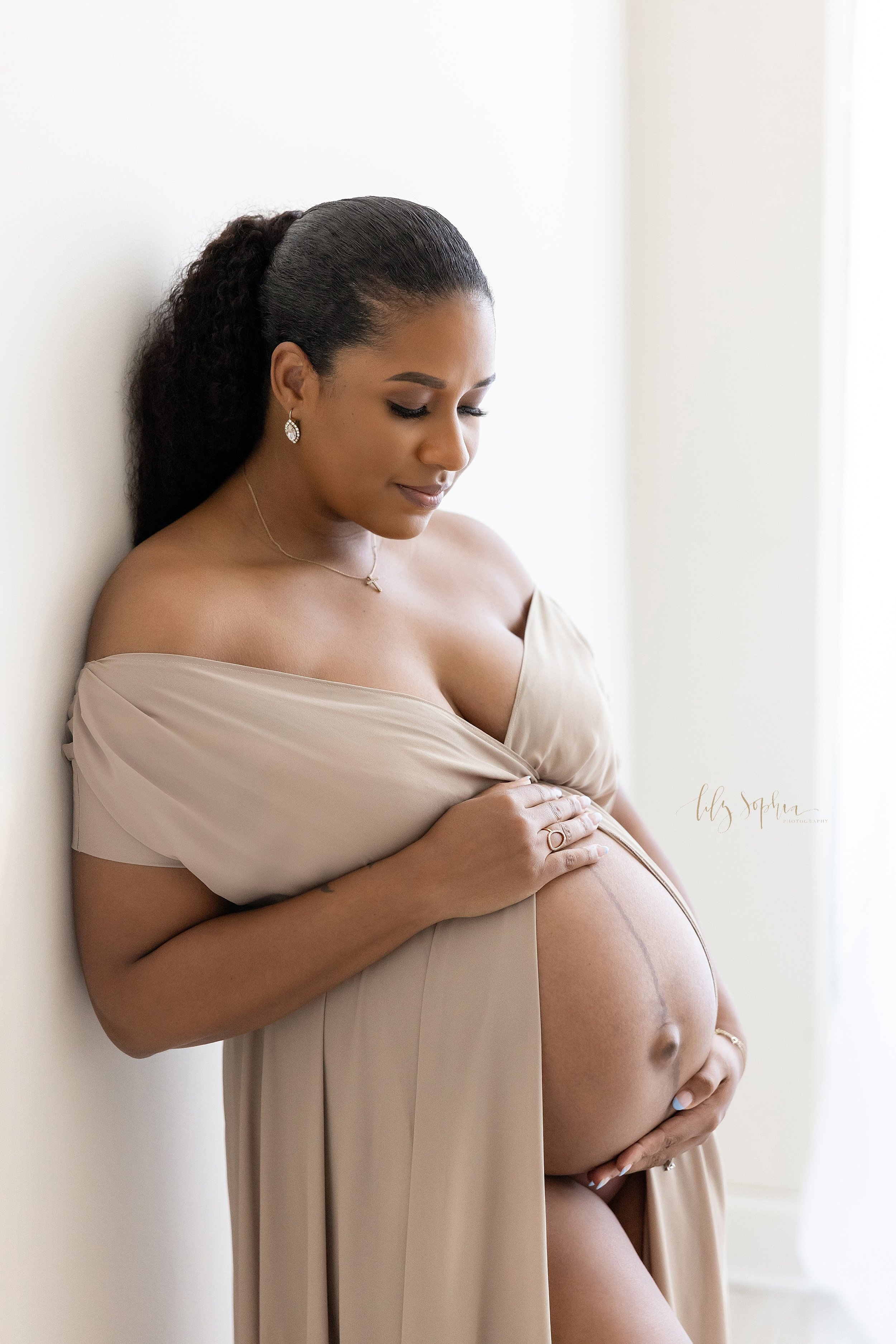  Maternity portrait of an African-American mother as she stands with her back against the wall of natural light studio wearing a split front gown to bare her belly with framing it with both her hands while she looks down and contemplates the birth of