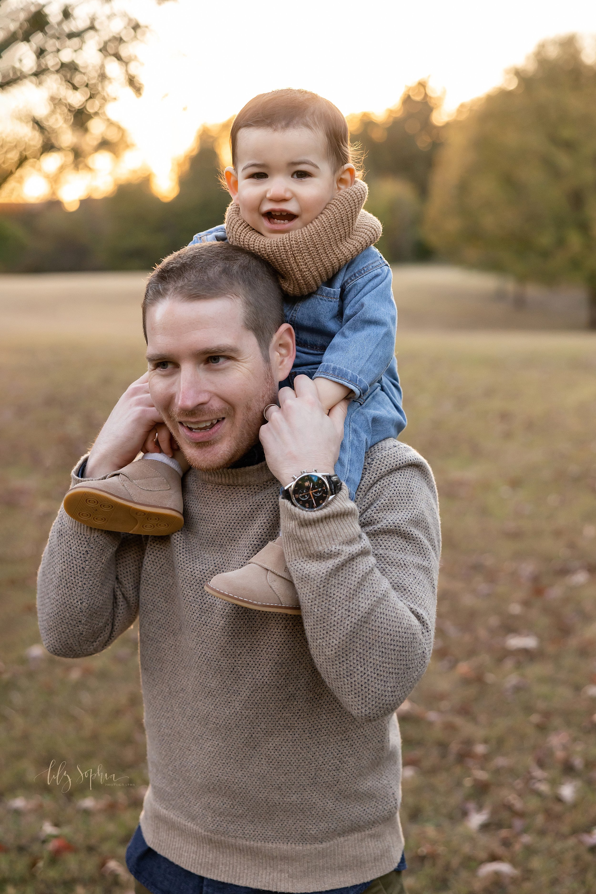  Family portrait of a father carrying his young son on his shoulders as the sun sets behind them during the fall season as he walks in an Atlanta park. 