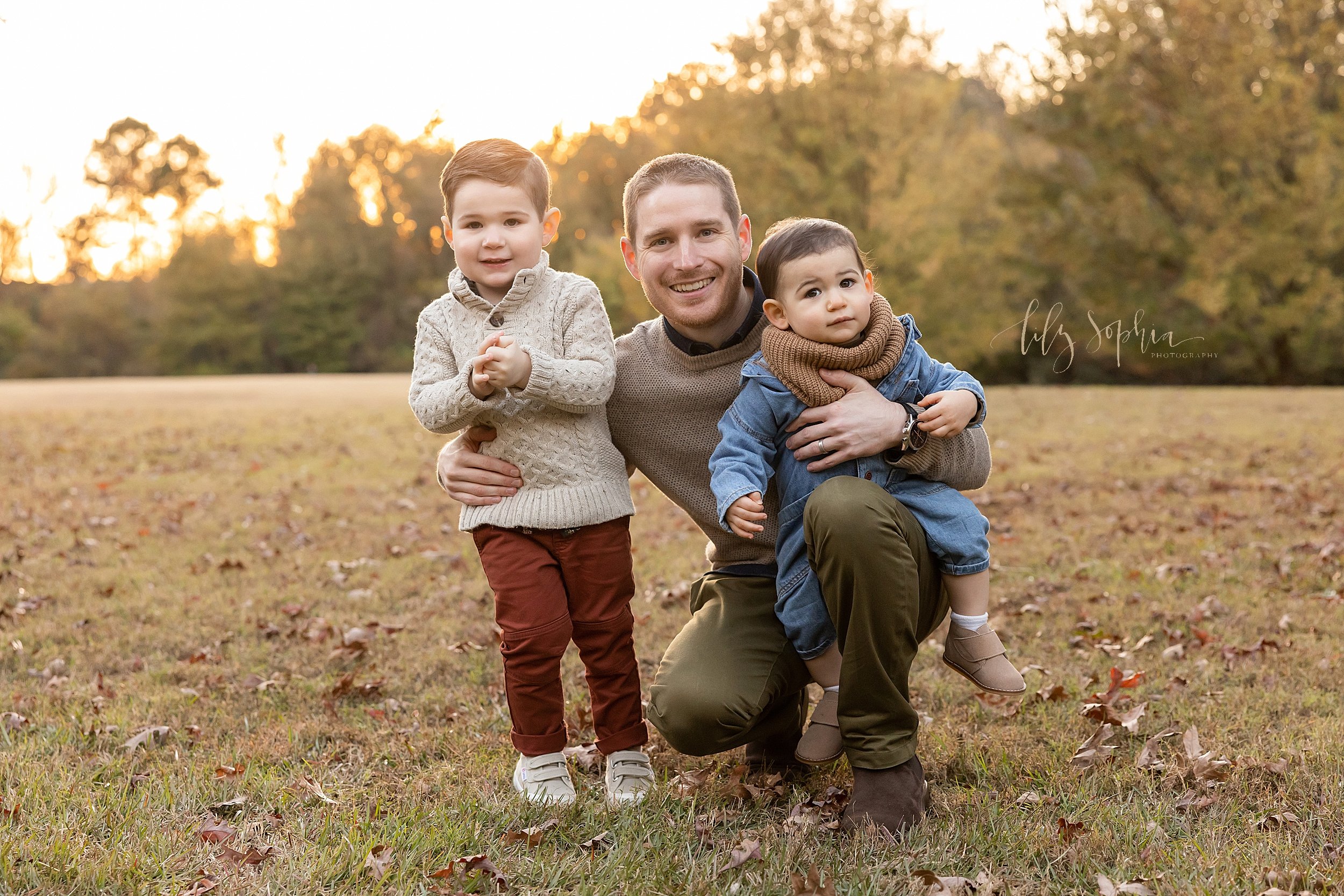  Family portrait of a father holding his youngest son on his knee as he squats next to his other son and holds him around the waist taken at sunset in an Atlanta park. 