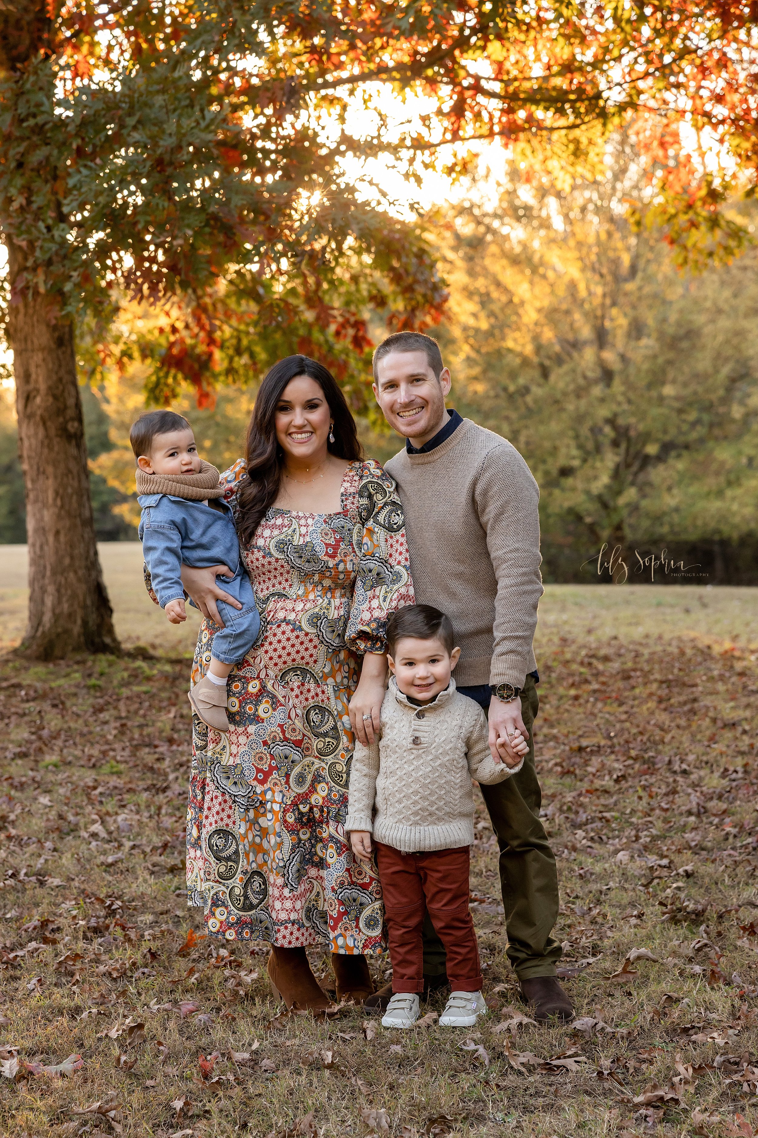  Family portrait of a mother holding her baby boy in her right arm as her husband stands beside her on her left side and her husband holds their toddler son’s hand with his left hand while the family is in an Atlanta park during autumn at sunset. 