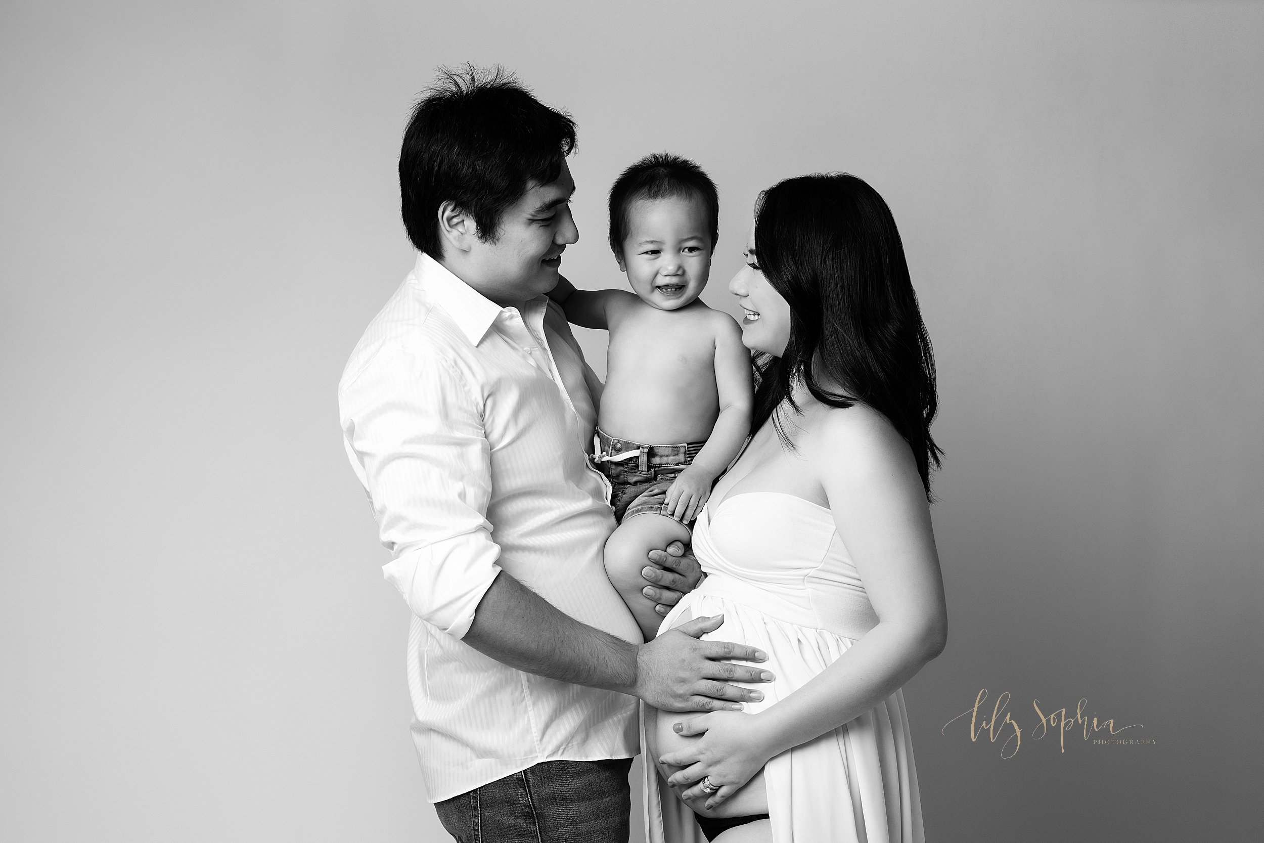 Modern black and white maternity session with an Asian family with dad holding their toddler son between him and his pregnant wife as she wears a strapless split front gown and bares her belly as the family talks with one another taken near Poncey H