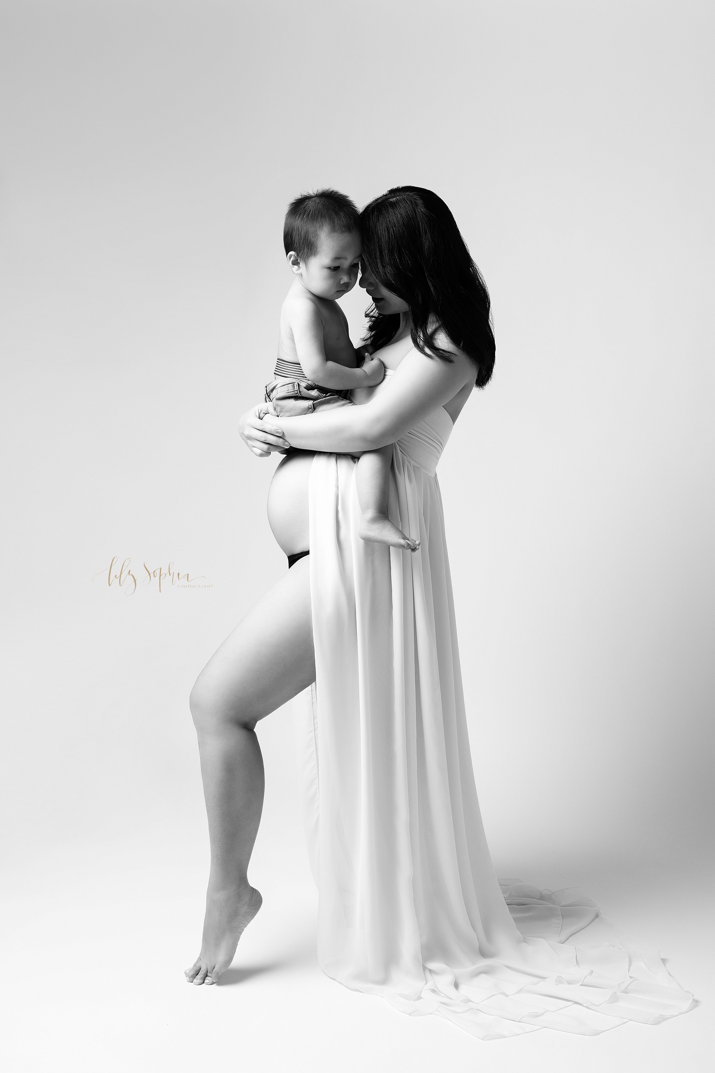  A modern maternity session with a expectant Asian mother as she stands holding her son wearing a split front full-length gown that bares her belly as she reveals her right leg while pointing her right toe taken in a studio in Ponce City Market in At