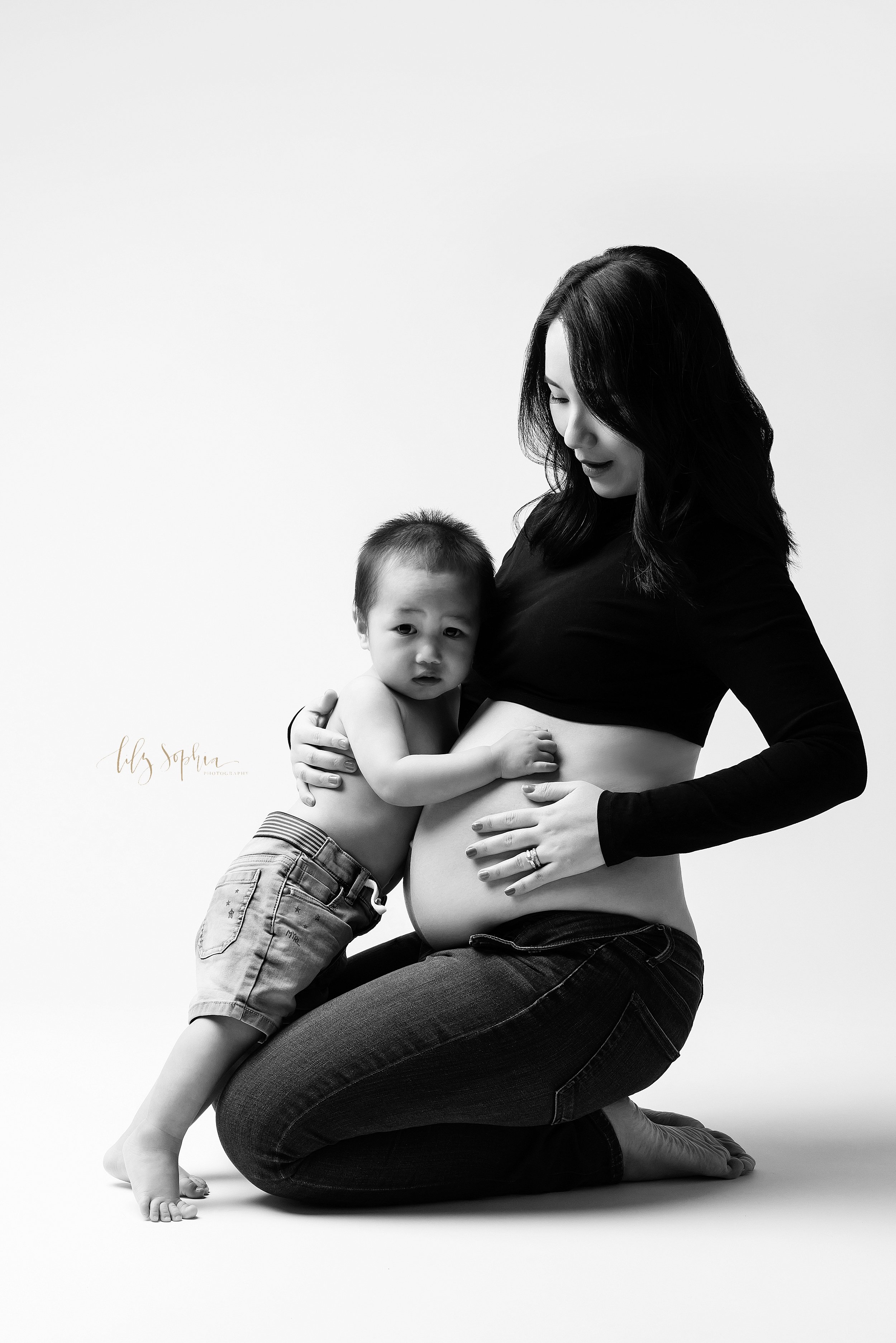  Modern maternity photo of a pregnant Asian woman kneeling on the floor of a studio with her belly bare and hugging her toddler son taken near Oakhurst in Atlanta. 