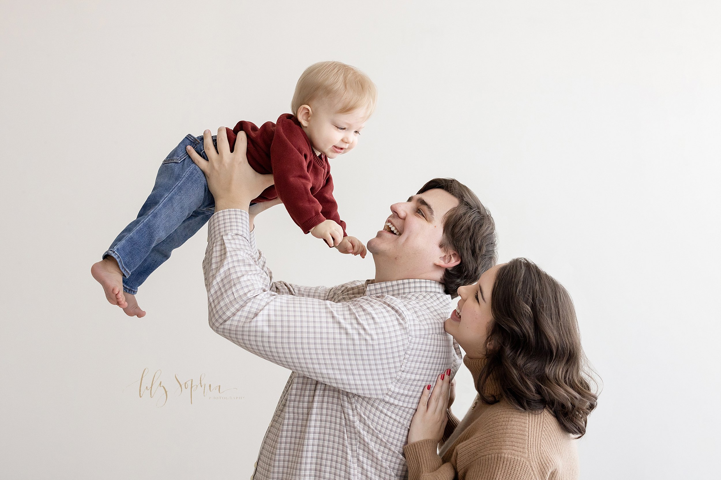  Family first birthday photo of a mother standing behind her husband as her husband lifts their one year son above his head taken near Morningside in Atlanta in a studio using natural light. 