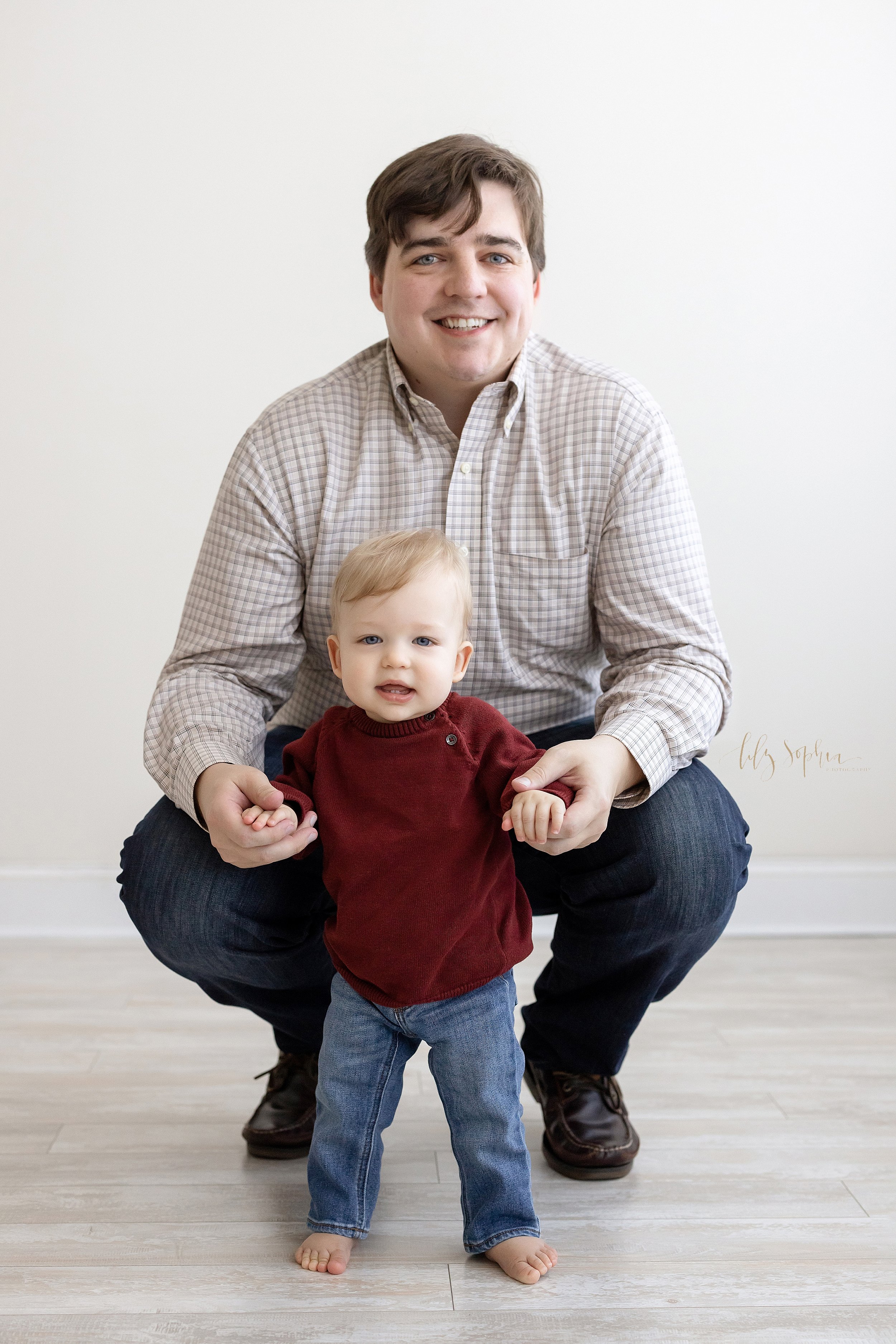  First birthday family photograph of a father squatting behind his one year old son and holding his son’s hands as his son stands in front of him taken in a natural light studio near Kirkwood in Atlanta, Georgia. 