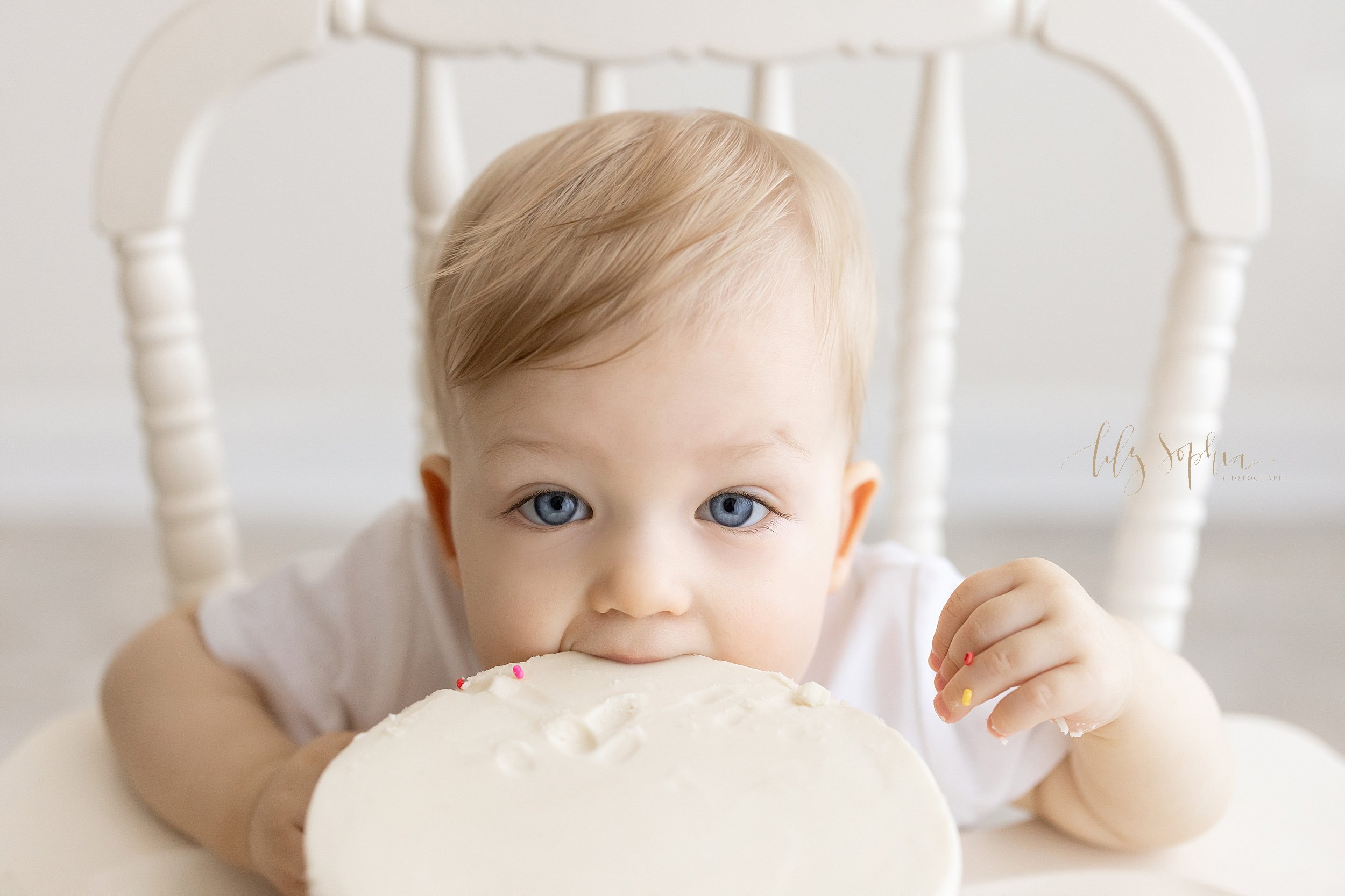  First birthday photo session of a one year old blue eyed boy as he sits in an antique high chair and bites into his smash cake taken in natural light in a studio near Buckhead in Atlanta, Georgia. 