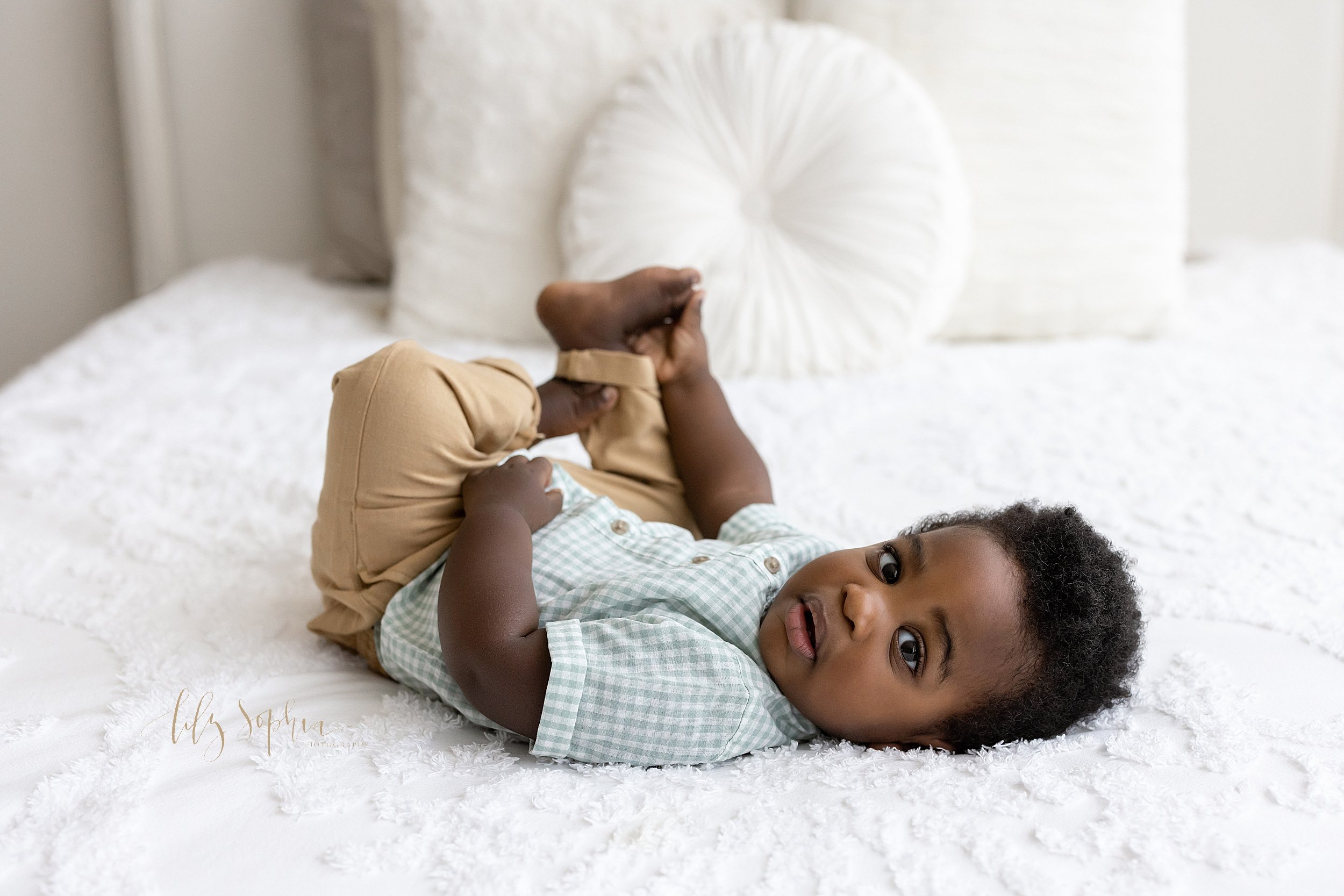  First birthday portrait of a black baby boy lying on his back on a bed and playing with his feet taken in a studio near Decatur in Atlanta, Georgia that uses natural lighting. 