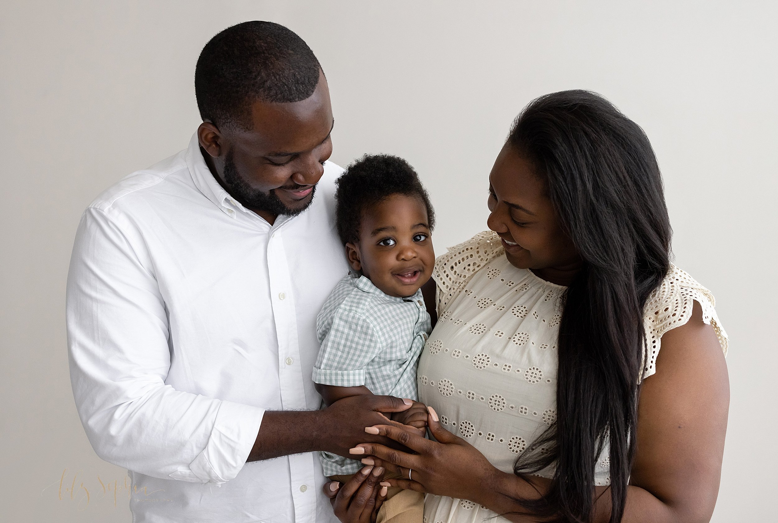  Family first birthday portrait of an African-American one year old boy being held by his mother with his father behind him as the parents smile and talk to their son taken in a natural light studio near Old Fourth Ward in Atlanta. 