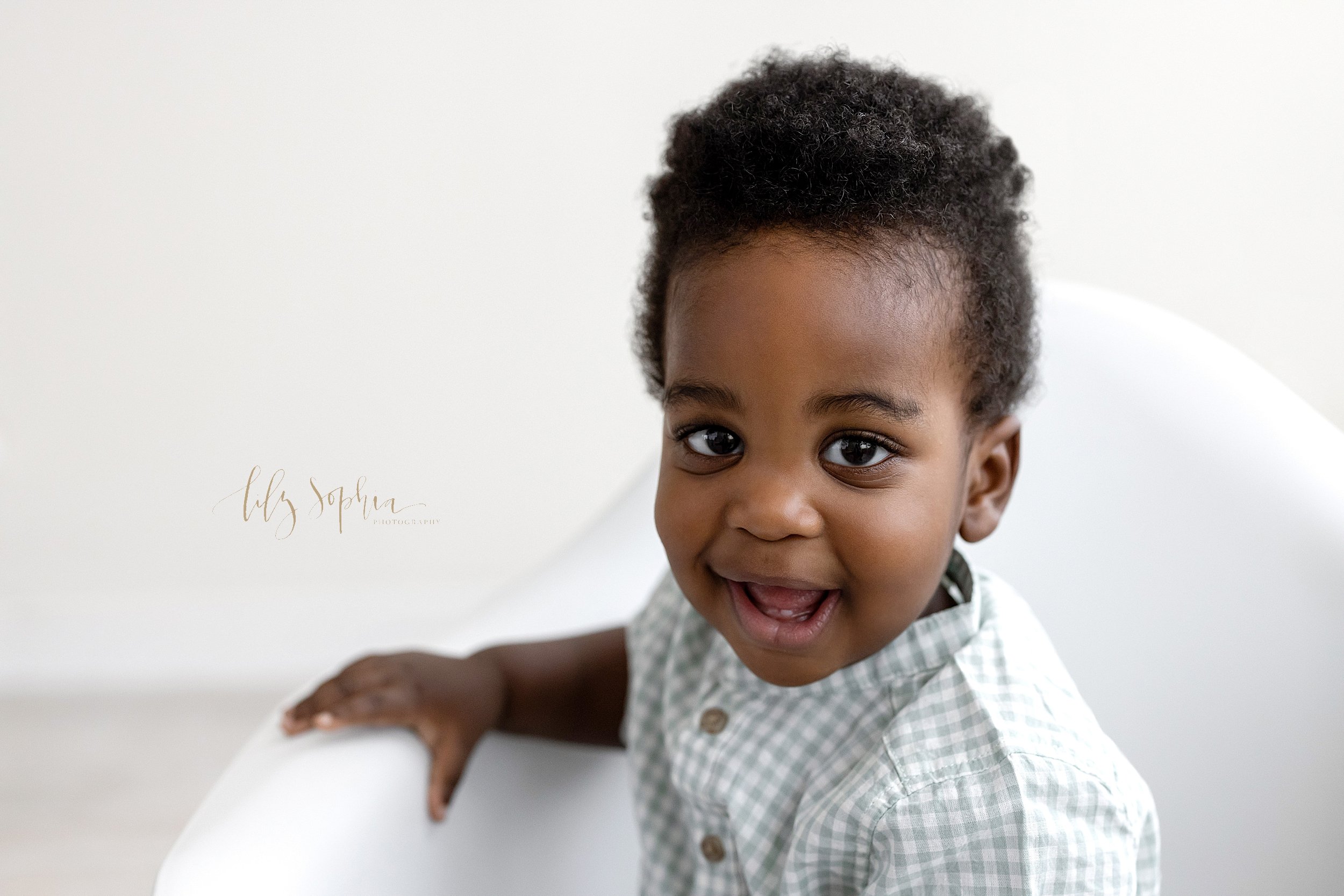  First birthday portrait of an African-American one year old boy as he sits in a white molded chair in front of a window streaming natural light and smiles to show his tiny teeth taken in a studio near Morningside in Atlanta, Georgia. 