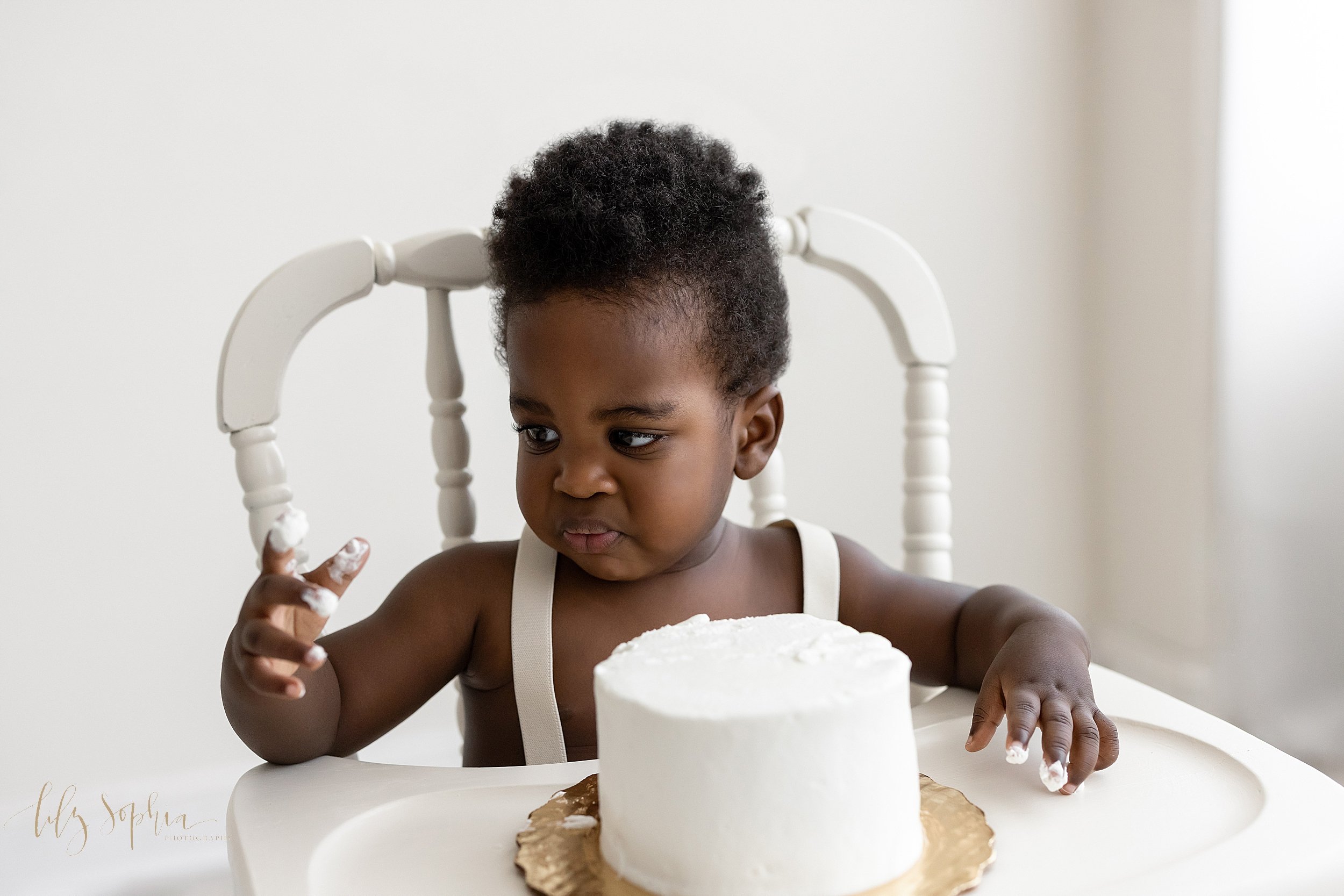  First birthday smash cake photograph of a black one year old boy as he sits in an antique high chair in front of a window streaming natural light and frowns about the icing that he has on his fingers taken in a studio in Ponce City Market. 