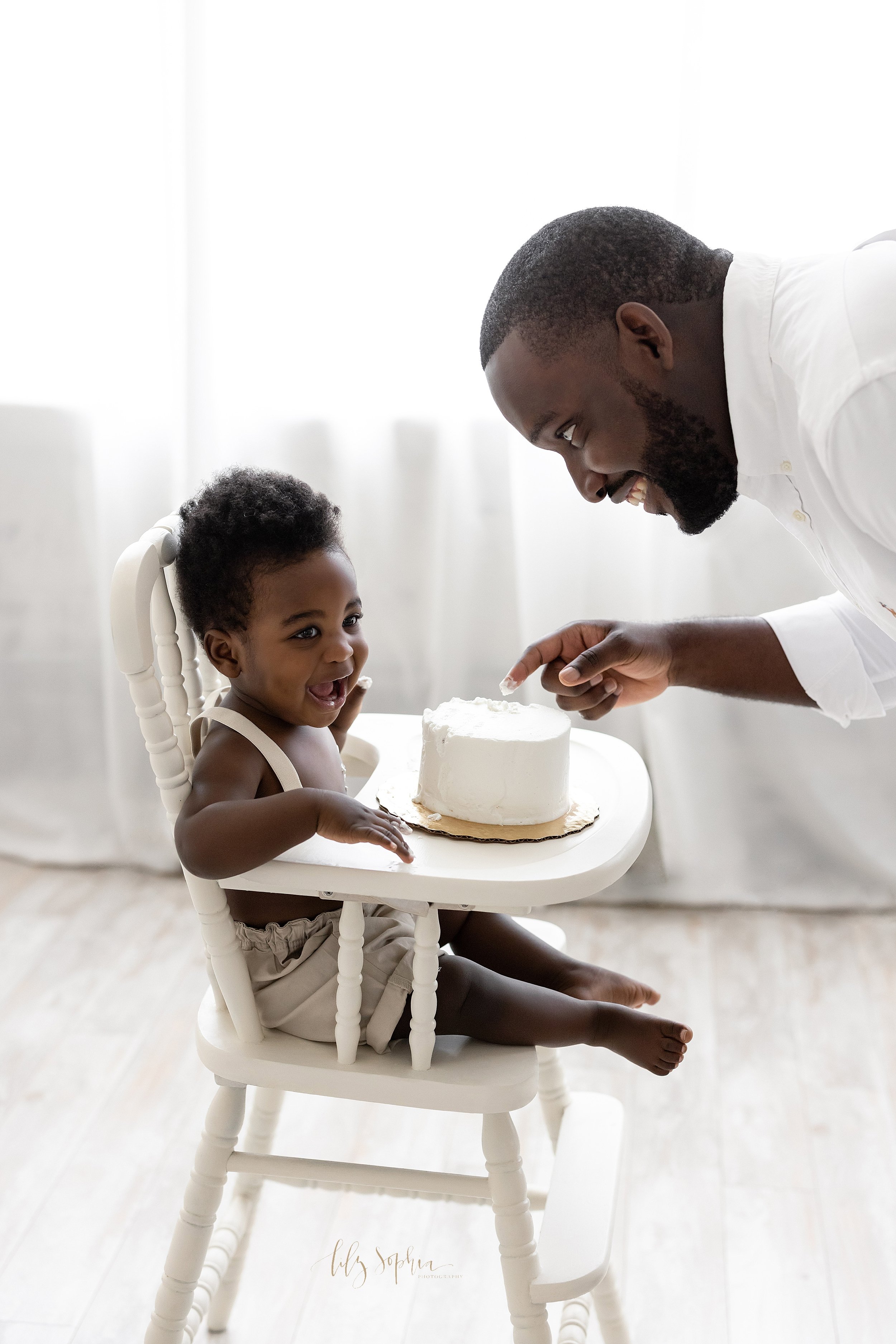  First birthday smash cake photograph of a one year old African-American boy as he sits in an antique high chair in front of a natural light window with his dad facing him with icing on the son’s and the dad’s hands as they both laugh taken near Smyr