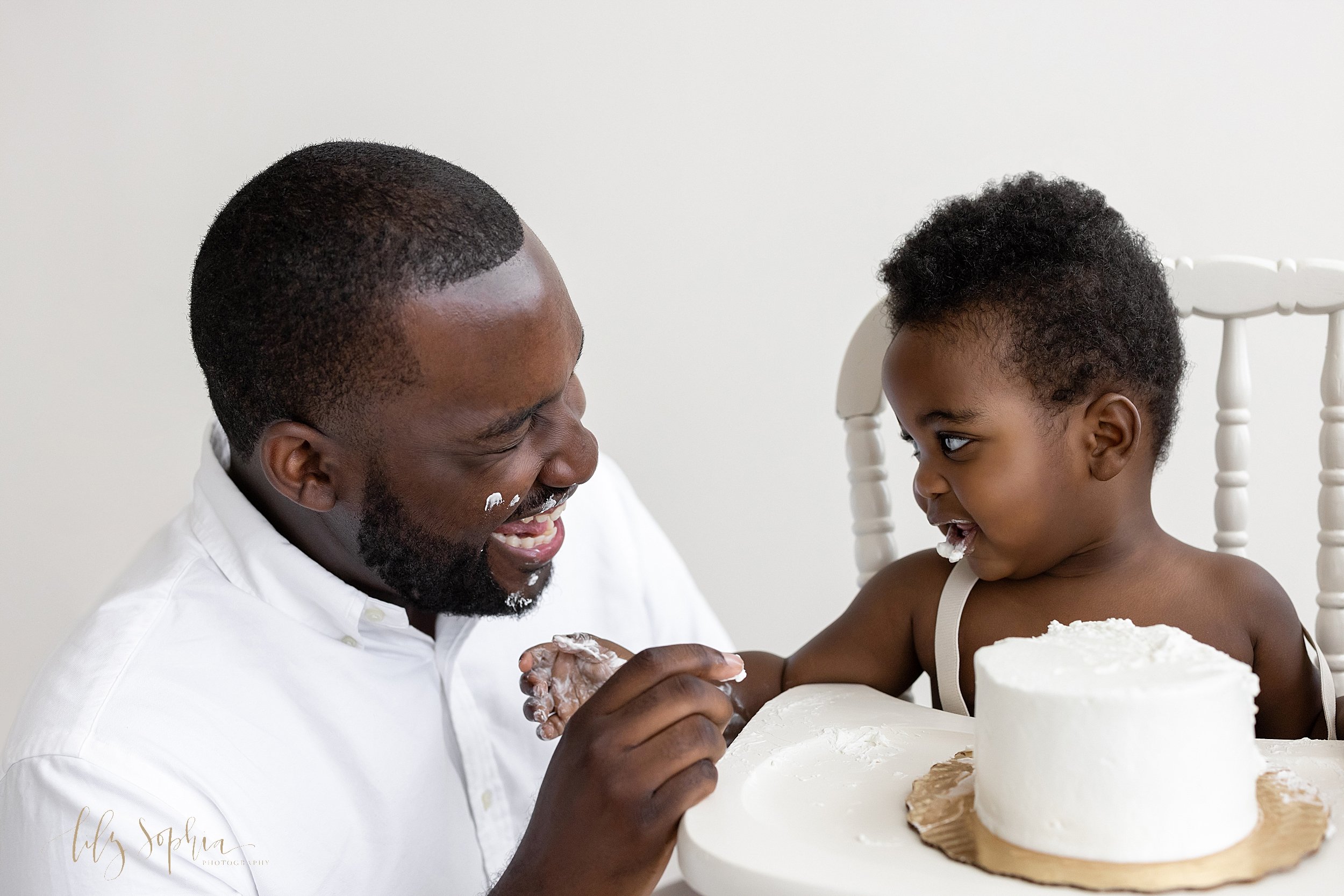  First birthday smash cake photo of a black father as he squats next to his son sitting in an antique high chair as his father laughs because the son has smeared icing on the father’s face and on his own taken in a natural light studio near Roswell i