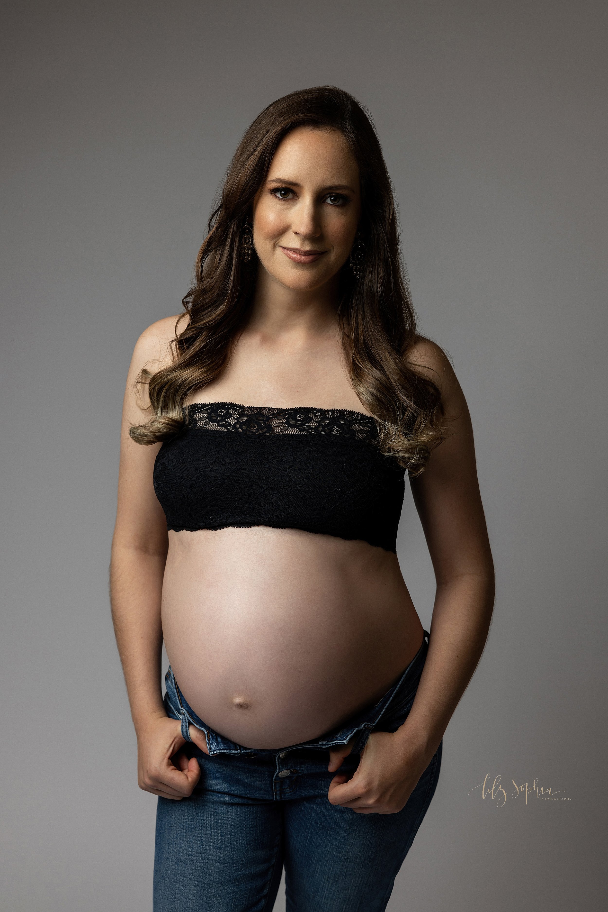  Modern maternity pictures of a pregnant woman as she stands wearing a black lace-edged bandeau bra and a pair of blue jeans with her bare belly shown as she hooks her fingers in the belt loop of her jeans taken in a studio near Morningside in Atlant